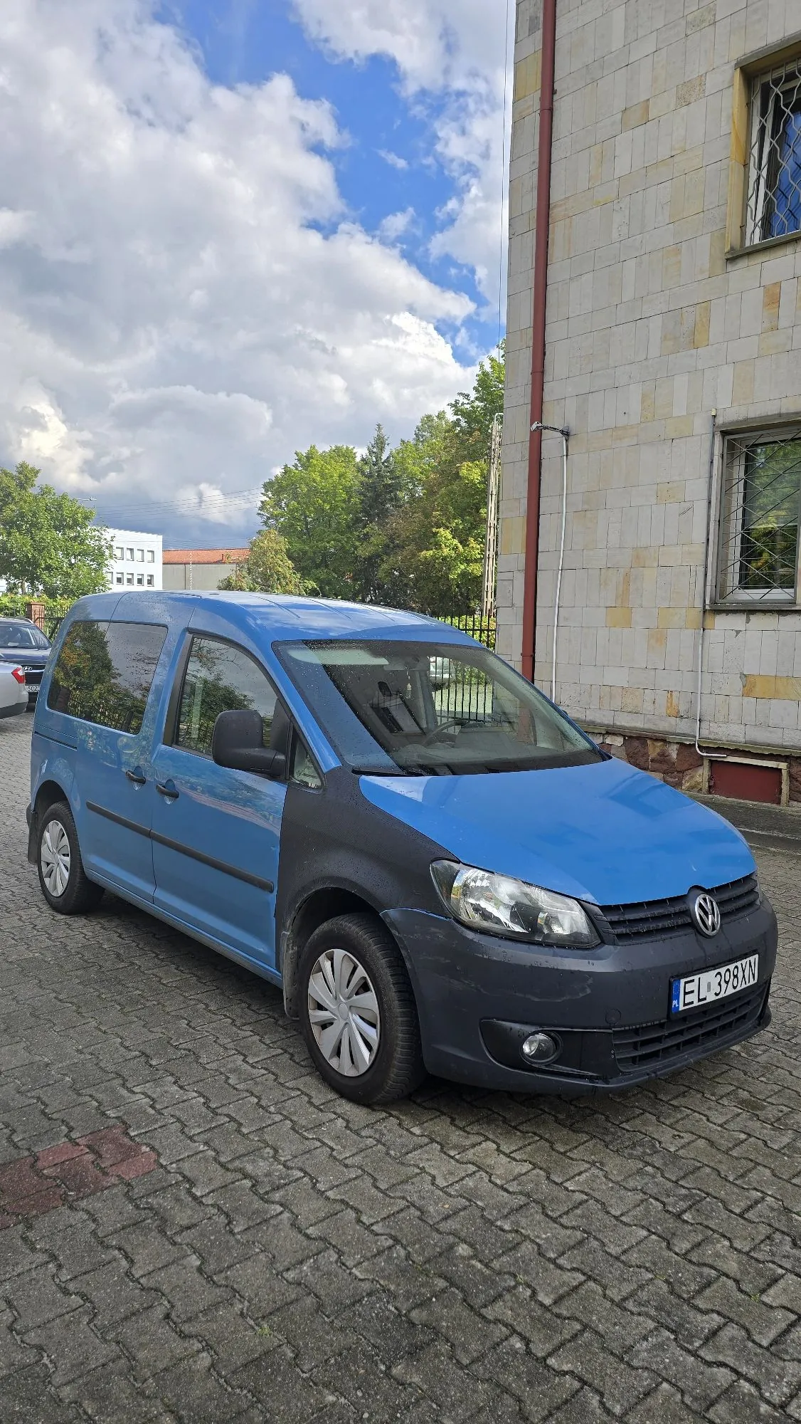 Niebieski samochód dostawczy marki Volkswagen zaparkowany na kostce brukowej obok jasnego, kafelkowanego budynku. Pojazd ma czarny zderzak przedni i czarne listwy boczne. Tablica rejestracyjna samochodu to EL 398XN. W tle widoczne są zielone drzewa oraz niebo z białymi chmurami.
