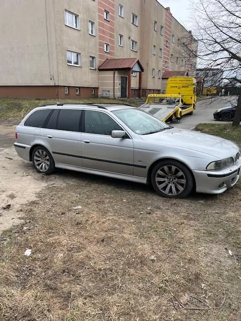 Srebrny samochód typu kombi zaparkowany na suchym trawniku. W tle żółty wóz holowniczy z platformą oraz wielopiętrowy blok mieszkalny z cegły i betonu. Widoczne są również suche drzewa i czarny samochód w oddali.