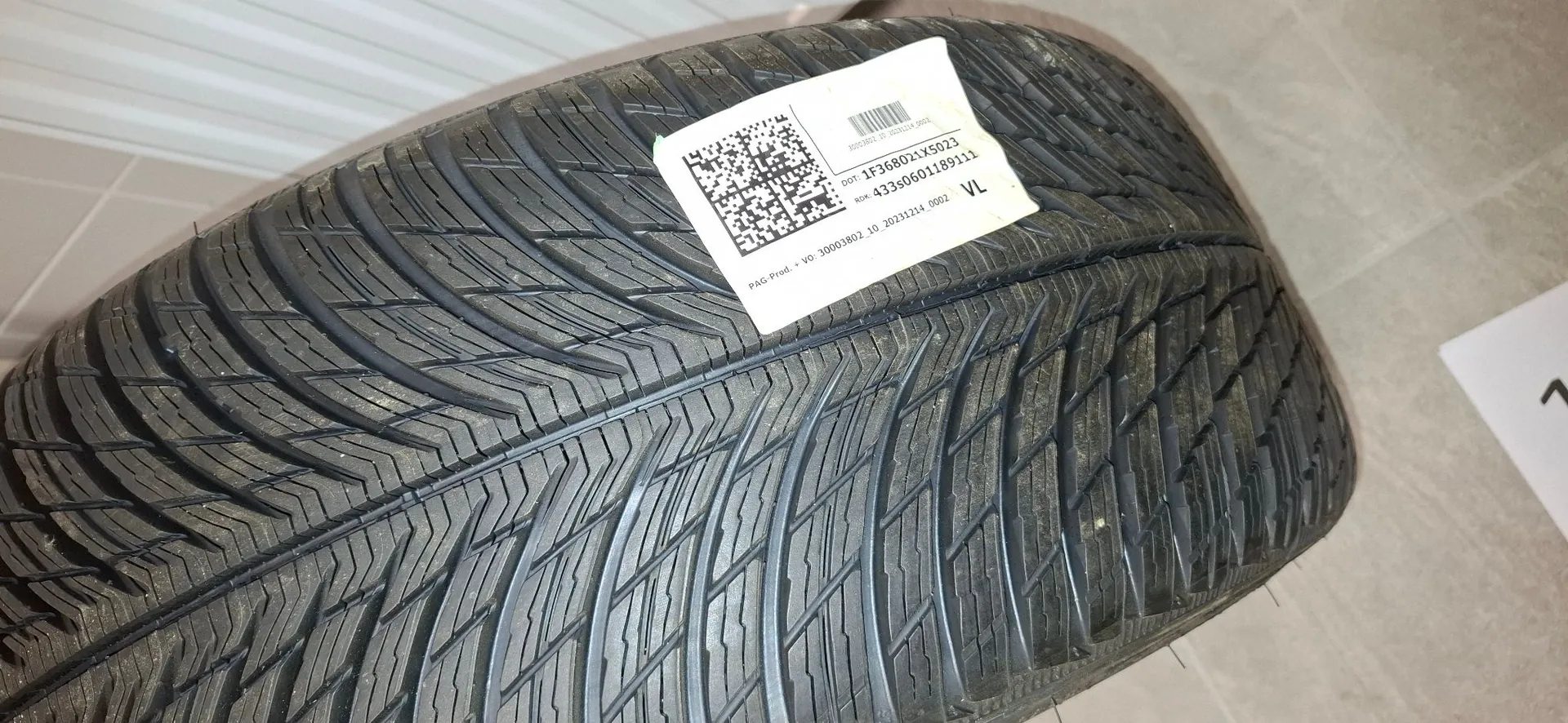 Zbliżenie na bieżnik opony samochodowej. Na boku opony naklejona jest biała etykieta z kodem QR oraz tekstem: 'DOT 1F368021X5023', 'KOD: 433s0601189111' oraz mniejszym drukiem na dole 'PAG Prod. + VO 30003802 10 20231214 0002 VL'.