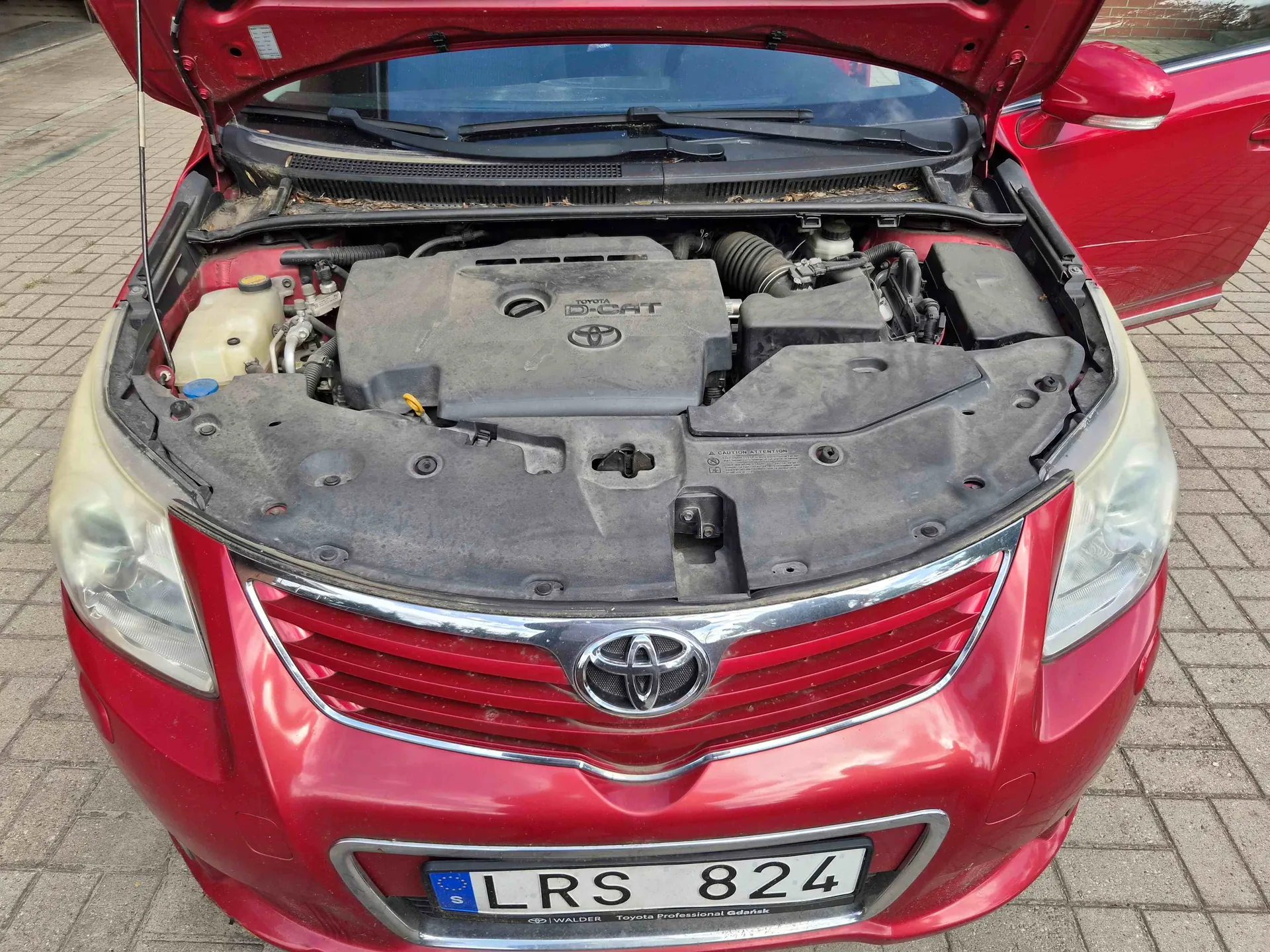 Zdjęcie przodu czerwonego samochodu marki Toyota z otwartą maską, ukazującą silnik. Na masce silnika widnieje napis 'D-CAT' oraz logo Toyoty. Samochód posiada polską tablicę rejestracyjną o numerze 'LRS 824'. Poniżej numeru rejestracyjnego znajduje się napis 'WALDER Toyota Professional Gdańsk'.