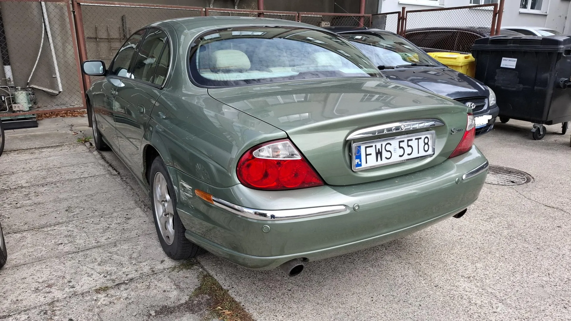 Tylny lewy trzy czwarte widok srebrno-zielonego samochodu marki Jaguar z polską tablicą rejestracyjną o numerze FWS 55T8. Samochód zaparkowany jest na betonowym podjeździe obok innego, ciemnego auta oraz czarnego kontenera na odpady.