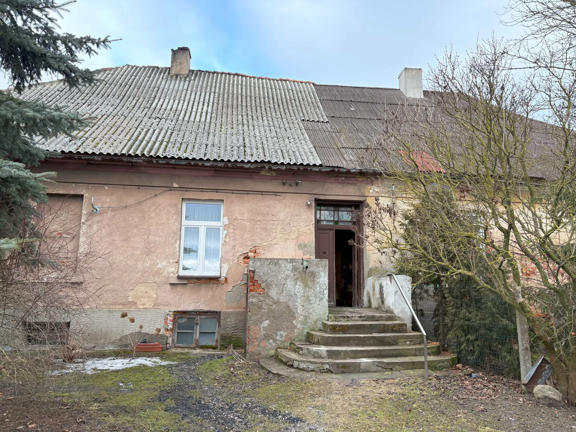 Stary, dwupiętrowy dom z uszkodzoną elewacją w kolorze bladego różu. Dach pokryty blachą falistą z dwoma kominami. Na parterze widoczne są okno z białą ramą, drzwi wejściowe z otwartym progiem oraz schody prowadzące do drzwi z metalową poręczą. Elewacja częściowo odsłania cegłę, a na ścianie znajdują się plamy i uszkodzenia. Wokół domu rosną drzewa bez liści i zielone krzewy, na ziemi widoczne są plamy trawy i żwiru. Niebo jest częściowo zachmurzone.