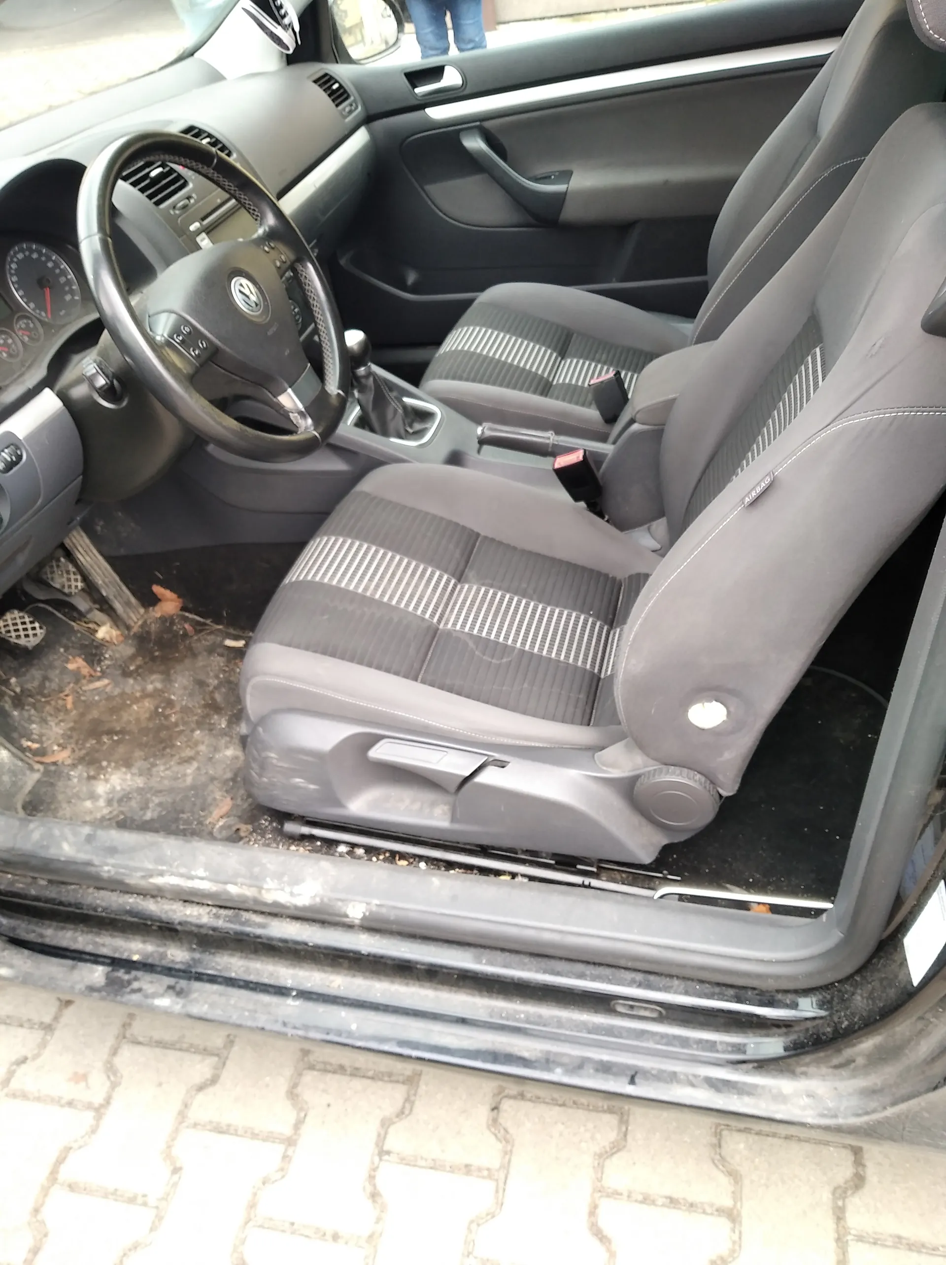 Wnętrze samochodu z widocznym kierownicą marki Volkswagen, szarymi siedzeniami z pionowymi prążkami, deską rozdzielczą z licznikami oraz brudną podłogą z liśćmi i śmieciem. Drzwi pasażera są otwarte, a na zewnątrz widoczna jest brukowana powierzchnia. W tle częściowo widoczne są nogi osoby w niebieskich spodniach.