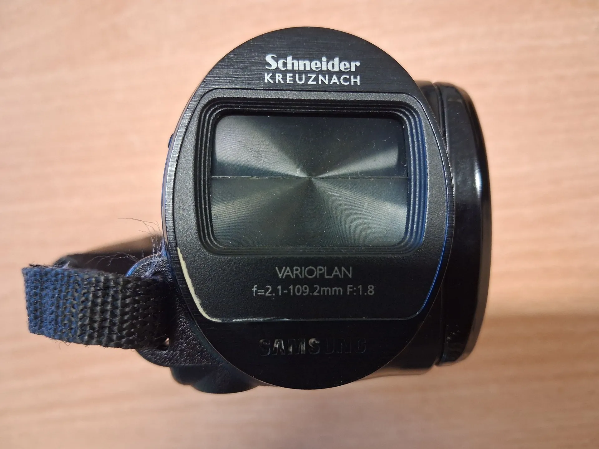 Zbliżenie na obiektyw aparatu marki Samsung z optyką Schneider Kreuznach. Na czarnej obudowie widnieją białe napisy: 'Schneider KREUZNACH' na górze oraz 'VARIOPLAN f=2.1-109.2mm F:1.8' poniżej soczewki. Na samym dole znajduje się wytłoczony napis 'SAMSUNG'. Do obudowy przymocowany jest czarny pasek z materiału.