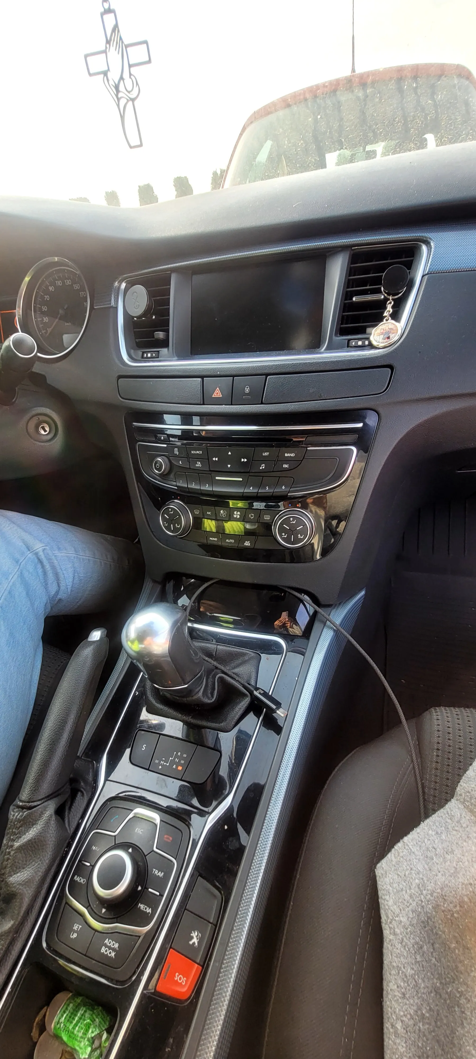 Wnętrze samochodu z widoczną konsolą środkową, prędkościomierzem po lewej stronie, centralnym ekranem, klimatyzacją z przyciskami i pokrętłami, dźwignią biegów z metalową gałką, kluczykami z krzyżem na lusterku wstecznym, nogami osoby w niebieskich dżinsach po lewej stronie oraz czerwonym przyciskiem SOS na konsoli. W przestrzeni na przedmioty widoczny zielony obiekt.