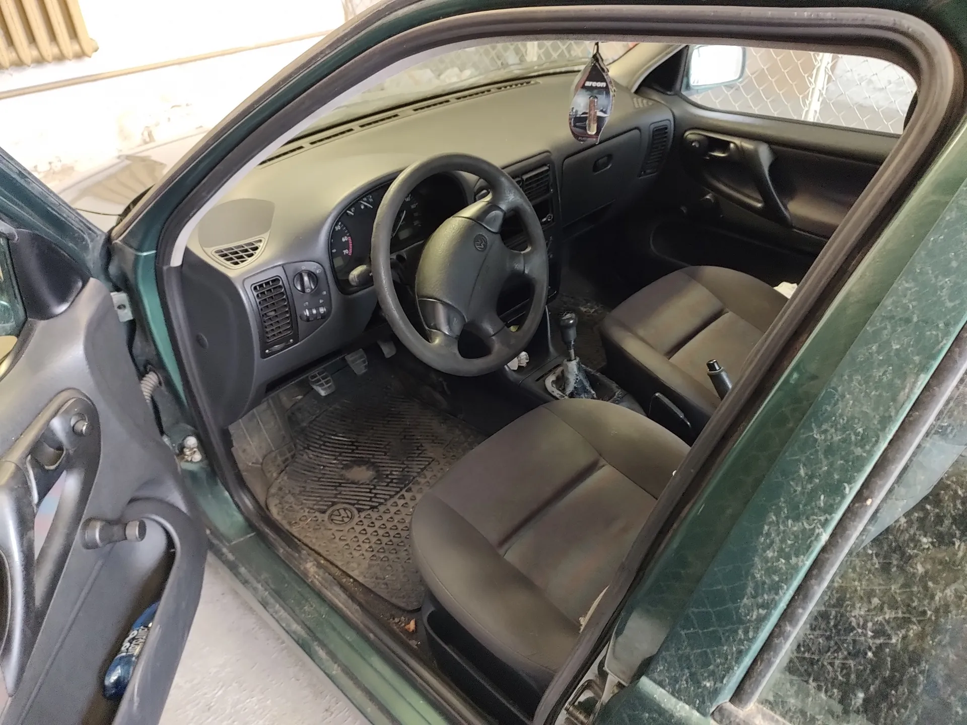Wnętrze samochodu z otwartą lewą przodem drzwiami. Widoczna kierownica z logo Volkswagen, deska rozdzielcza z licznikami, brązowe siedzenia, brudne dywaniki podłogowe z logo VW oraz dźwignia zmiany biegów. Zewnętrzna część drzwi i karoseria samochodu są zielone. W tle za szybą widać siatkę ogrodzeniową i fragment innego pojazdu.