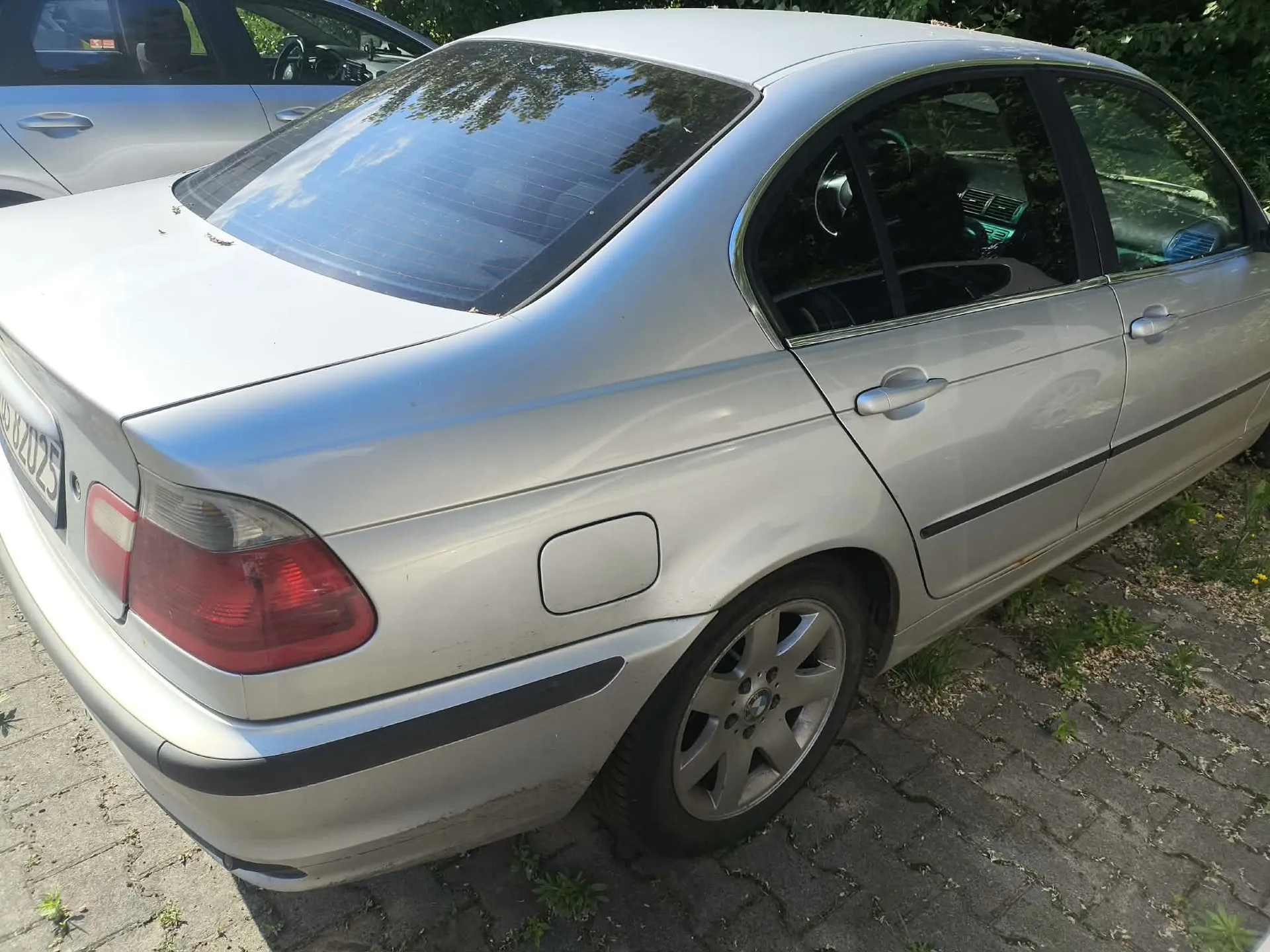 Srebrny samochód osobowy marki BMW serii 3 (E46) zaparkowany na kostce brukowej. Zdjęcie przedstawia tył i prawy bok pojazdu. Widoczne są tylne światła, klapka wlewu paliwa oraz felgi aluminiowe. W tle widać fragment innego srebrnego samochodu oraz zielone krzewy.