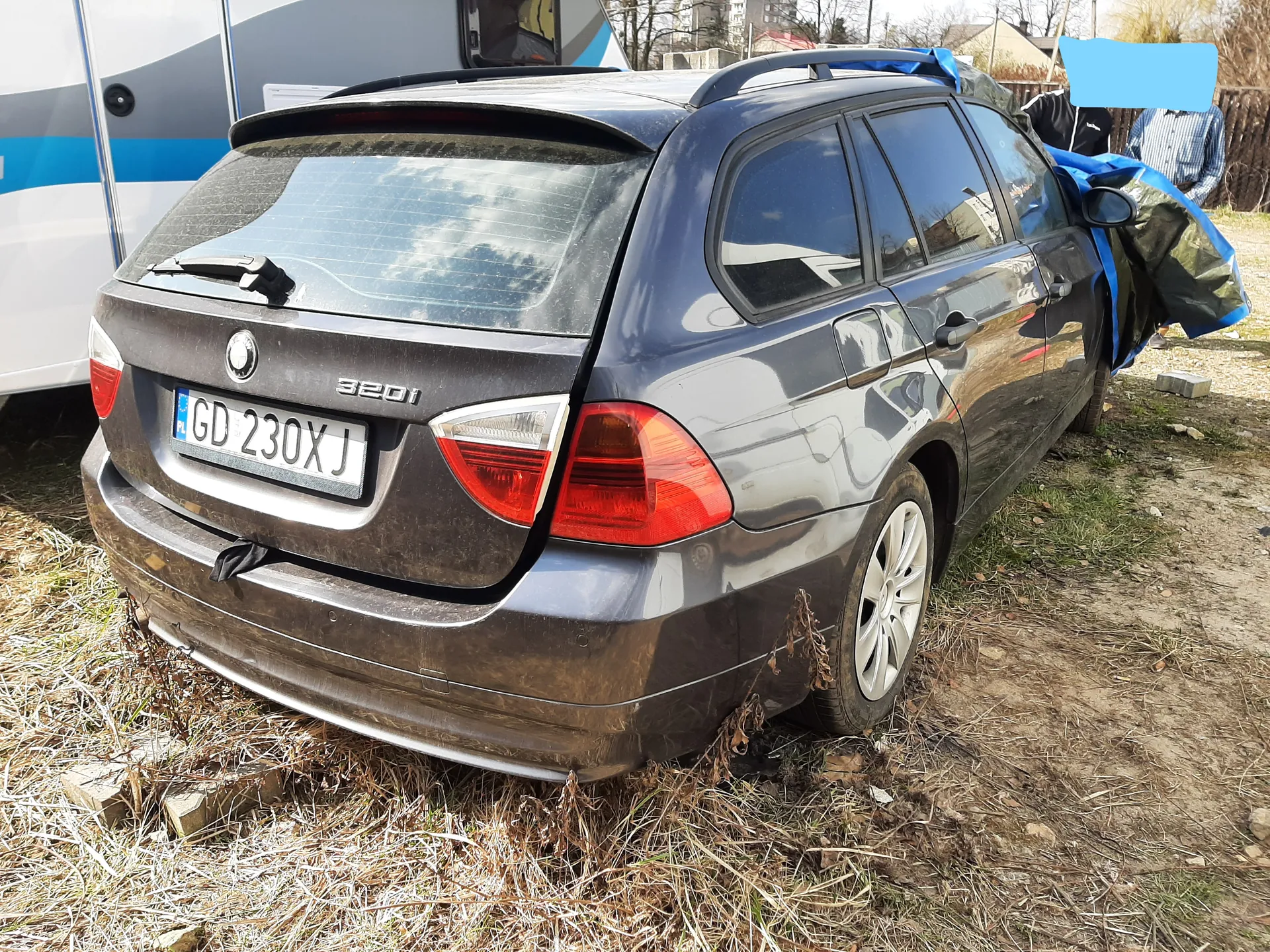 Ciemnoszary samochód osobowy typu kombi marki BMW 320i z tablicą rejestracyjną GD 230XJ (z polską flagą po lewej stronie). Samochód stoi na trawiastym i ziemnym terenie, z prawej strony częściowo przykryty niebiesko-czarną folią. W tle widoczny biało-niebieski przyczepa lub pojazd rekreacyjny, drzewa, budynki oraz osoba w niebieskiej koszuli. Na tylnym szybie refleksy chmur, a na podłodze sucha trawa i kilka cegieł.