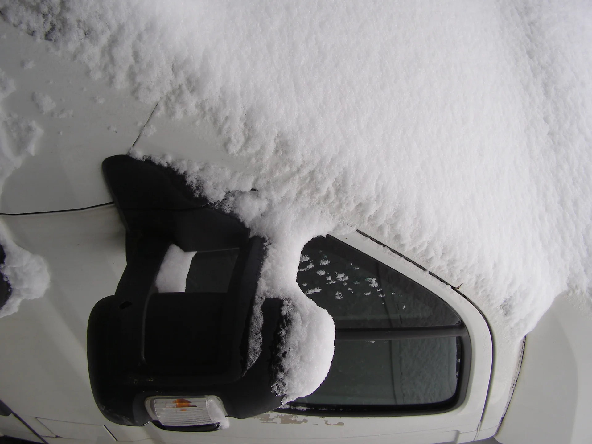 Biały samochód pokryty grubą warstwą śniegu, szczególnie na dachu i lusterku bocznym. Czarne lusterko z widocznym żółtym kierunkowskazem, częściowo zakryte śniegiem. Na szybie bocznej śnieżne płatki, a na karoserii ślady brudu. Tło zasłonięte śniegiem.