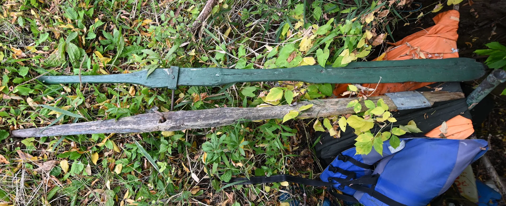 Zdjęcie przedstawiające przedmioty leżące na ziemi wśród zielonej i żółtej roślinności. Widoczny jest długi, zielony, drewniany przedmiot przypominający miecz lub replikę broni, oraz obok niego szary, zwietrzały kawałek drewna. W prawym dolnym rogu znajduje się niebieski plecak oraz fragmenty pomarańczowego materiału.
