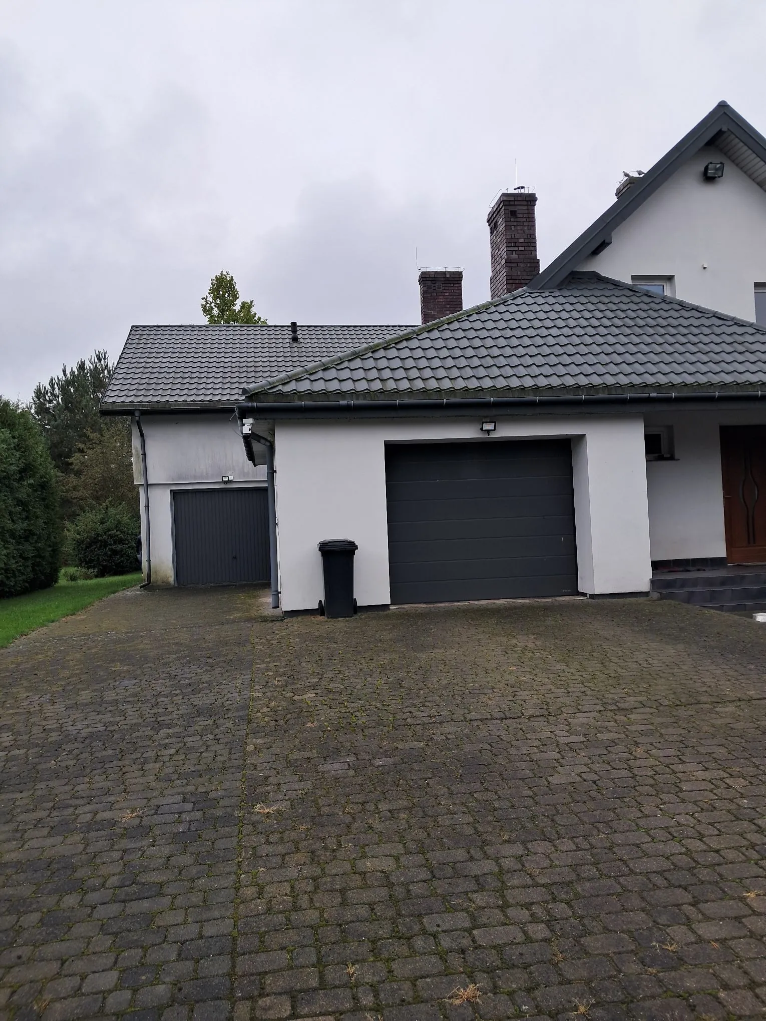 Zdjęcie domu jednorodzinnego z białymi ścianami i ciemnoszarym, spadzistym dachem z dachówki. Widoczne są dwa garaże z ciemnoszarymi bramami oraz czarny kosz na śmieci stojący obok jednego z nich. Przed domem znajduje się podjazd wyłożony szarą kostką brukową. W tle widać dwa kominy z czerwonej cegły oraz zielone drzewa pod zachmurzonym niebem.