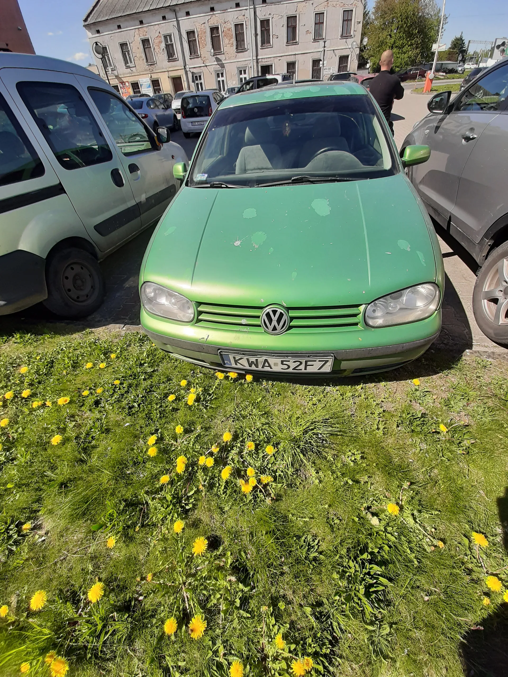 Zielony samochód marki Volkswagen z tablicą rejestracyjną KWA 52F7 zaparkowany na poboczu. Po prawej stronie obrazu zielona trawa z żółtymi kwiatami (mniszkiem lekarskim). W tle widoczne są inne pojazdy, w tym szary samochód oraz budynek z wieloma oknami. Na lewo od budynku stoi osoba. Na przedniej szybie zielonego samochodu widoczne są ślady uszkodzeń lakieru.