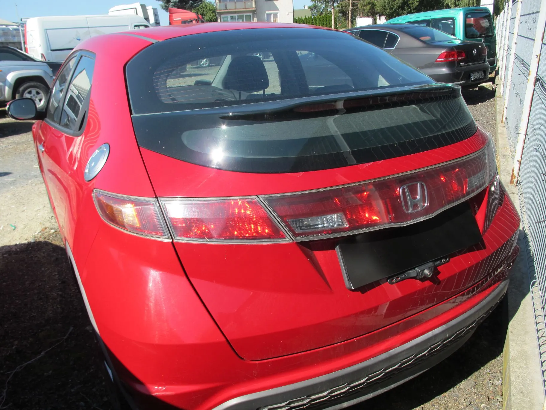 Tylny widok czerwonego samochodu marki Honda Civic zaparkowanego na zewnątrz. Widoczna jest tylna szyba z ciemnym spoilerem, logo Hondy na klapie bagażnika oraz tylne lampy. W tle znajdują się inne pojazdy, w tym biały furgon i szary samochód osobowy, oraz fragment ogrodzenia z siatki.