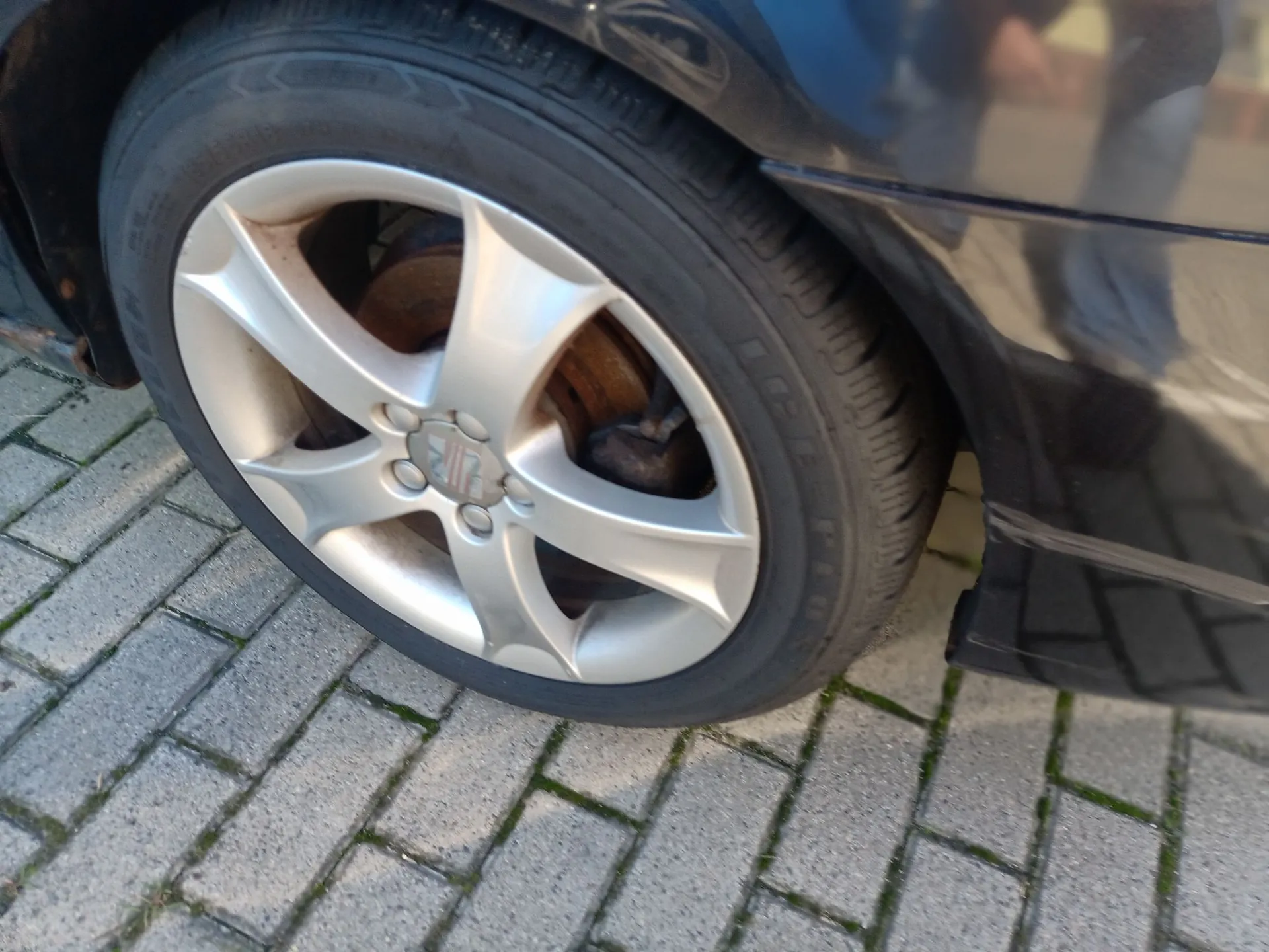 Zbliżenie na przednie koło samochodu w kolorze czarnym. Widoczna jest srebrna felga aluminiowa z pięcioma szerokimi ramionami i logo marki Suzuki w centrum. Opona jest zamontowana na feldze, a w tle widać fragment nadwozia auta oraz szarą kostkę brukową na ziemi.