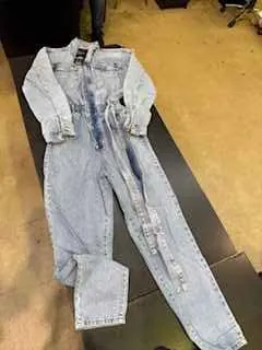 Jasnoniebieski dżinsowy kombinezon leżący na ciemnej powierzchni. Kombinezon ma długie rękawy, zamek na przodzie oraz przetarcia na nogawkach. Tło stanowi betonowa podłoga z widocznymi fragmentami mebli w kątach.