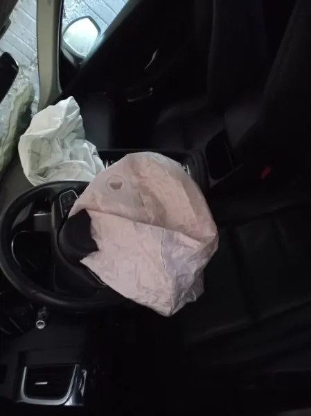 Wnętrze samochodu z rozwiniętymi poduszkami powietrznymi po stronie kierowcy i pasażera. Widoczne czarne siedzenia, kierownica oraz fragment ściany z kamienia za oknem. Wnętrze jest ciemne, a poduszki powietrzne są białe i zmięte.