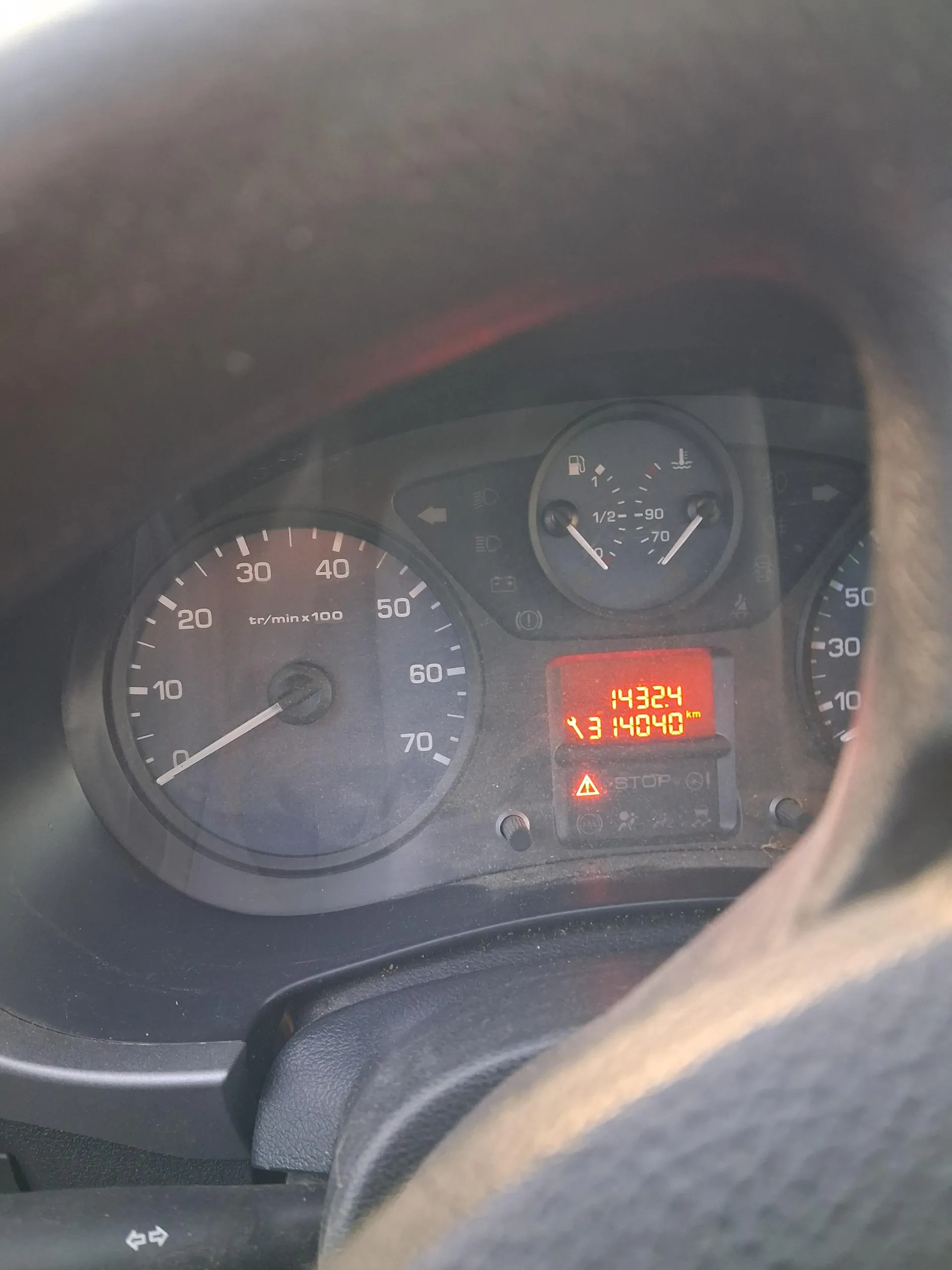 Zbliżenie na deski rozdzielcze samochodu. Po lewej stronie wskaźnik obrotomierza z oznaczeniem 'tr/min x100' i zakresem od 0 do 70. W centrum wyświetlacz z czerwonymi cyframi '14324' oraz '13 14040 km', poniżej czerwony trójkąt ostrzegawczy z napisem 'STOP'. W tle widoczne wskaźniki paliwa i temperatury chłodniczego z zakresem '1/2' do '90'. Fragment kierownicy widoczny u góry i dołu zdjęcia.