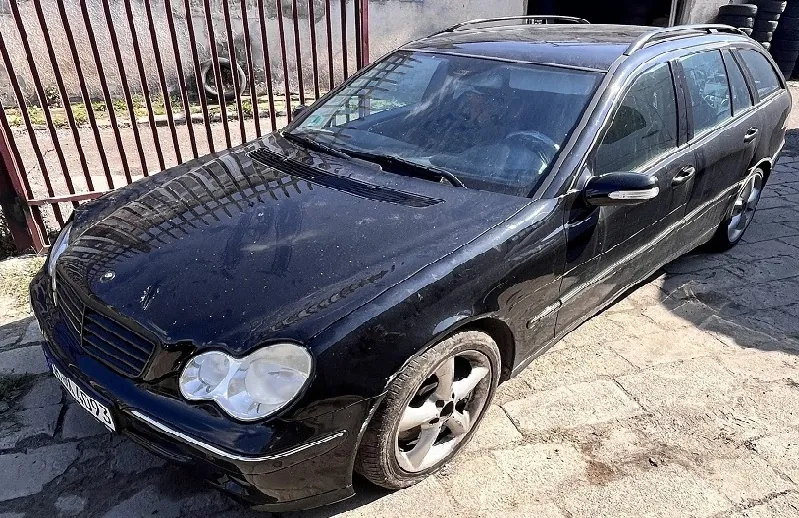 Czarny samochód osobowy w nadwoziu kombi, zaparkowany na brukowanej nawierzchni przed metalowym ogrodzeniem. Pojazd jest widoczny z przodu i z boku, ma srebrne felgi aluminiowe i widoczną tablicę rejestracyjną z przodu.