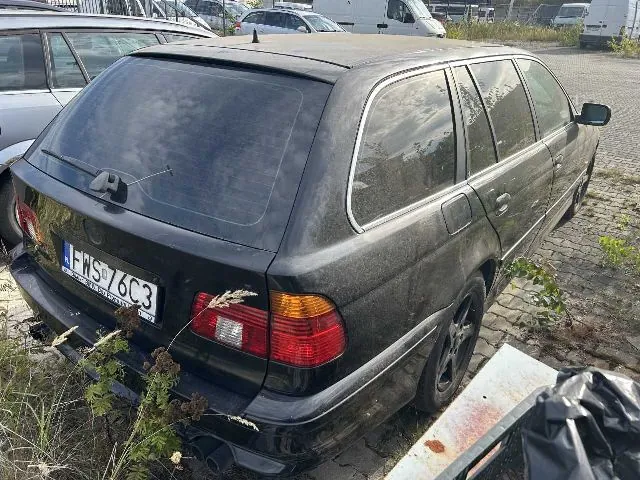 Czarny samochód osobowy typu kombi zaparkowany na kostce brukowej, widok z tyłu pod kątem. Pojazd jest zakurzony i posiada polską tablicę rejestracyjną o numerze FWS 76C3. W tle widoczne są inne zaparkowane samochody oraz biały furgon.