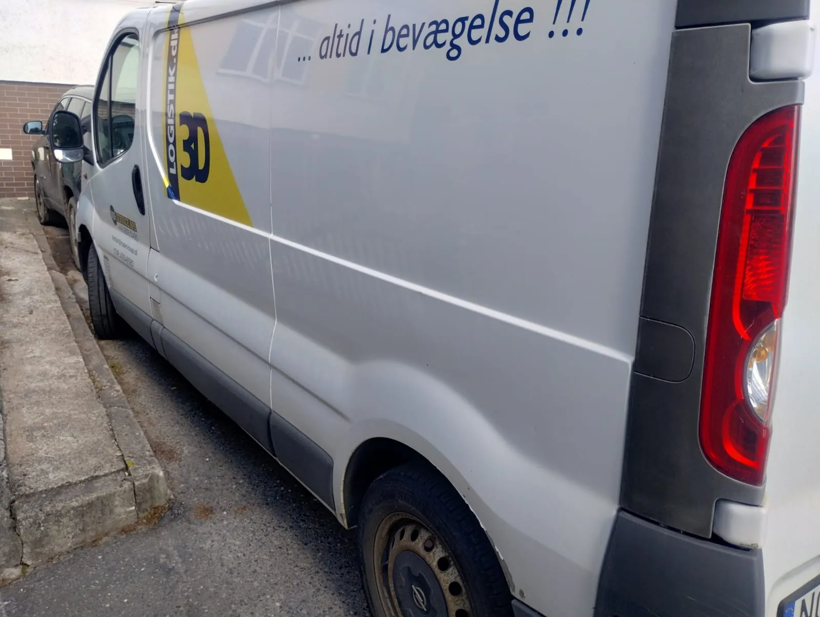 Srebrny furgon zaparkowany przy chodniku. Na boku widoczny żółto-niebieski napis 'LOGISTIK' oraz '3D', poniżej napis w języku duńskim '... altid i bevægelse !!!'. W tle czarny samochód i ceglana ściana. Widoczne czerwone światło tylnego reflektora.
