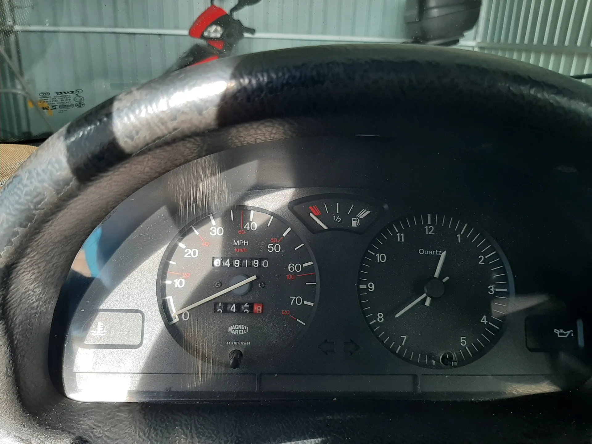 Zbliżenie na deski rozdzielcze samochodu. Widoczny czarny kierownica z metalowymi elementami, wskaźnik prędkości z oznaczeniami MPH i km/h (wskazówka na 0), licznik przebiegu 049190, mały wyświetlacz z cyframi 14 i 6, zegar z napisem Quartz pokazujący godzinę około 1:38, wskaźnik paliwa powyżej połowy. W tle przez przednią szybę widoczny czerwony skuter i metalowa ogrodzenie. Na desce rozdzielczej widoczne symbole, w tym napis Magneti Marelli.