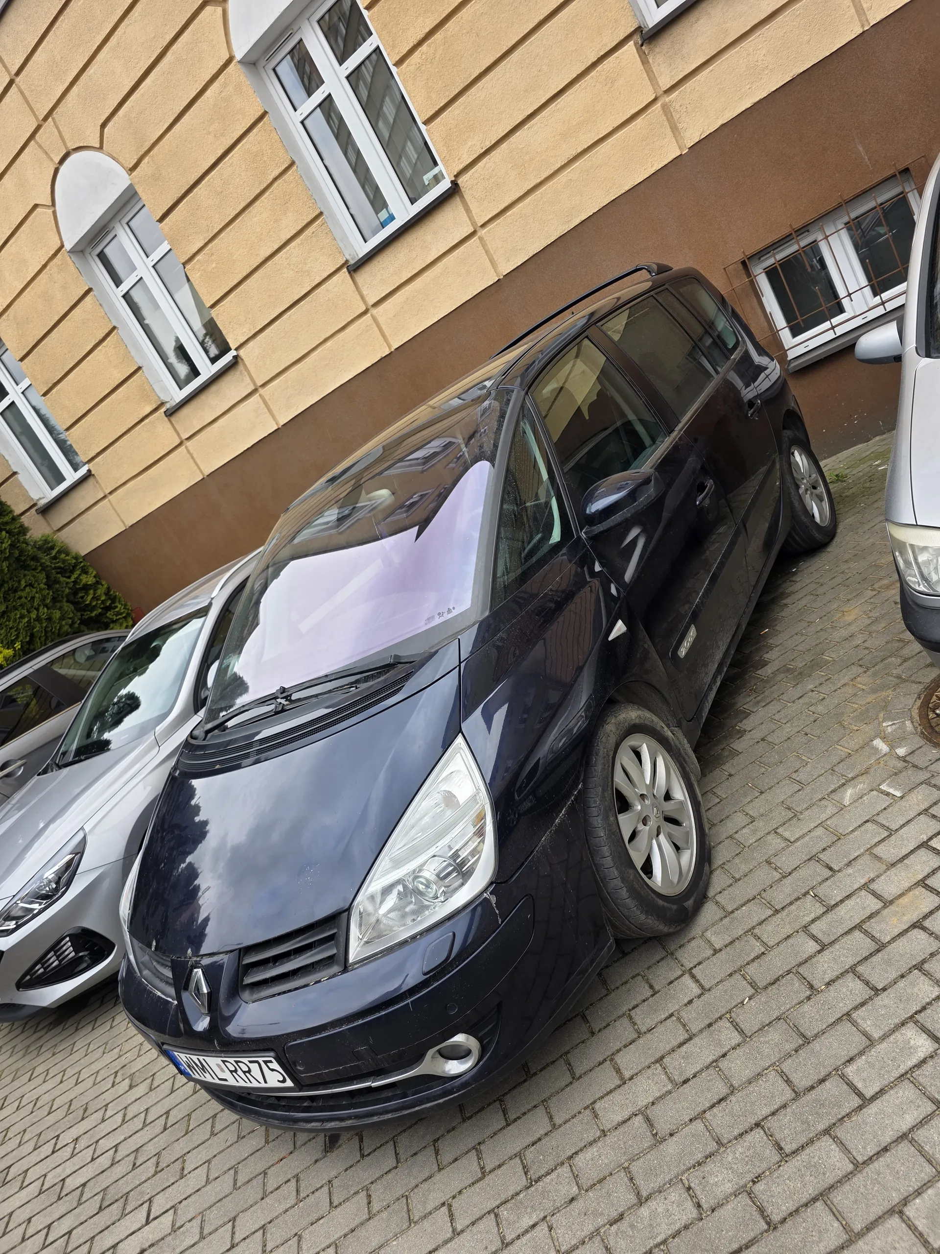 Czarny samochód zaparkowany na brukowanej nawierzchni z tablicą rejestracyjną WML R75. Obok czarnego samochodu znajduje się srebrny samochód. W tle budynki z beżowymi ścianami i białymi oknami. Część innego srebrnego samochodu widoczna w górnym lewym rogu.