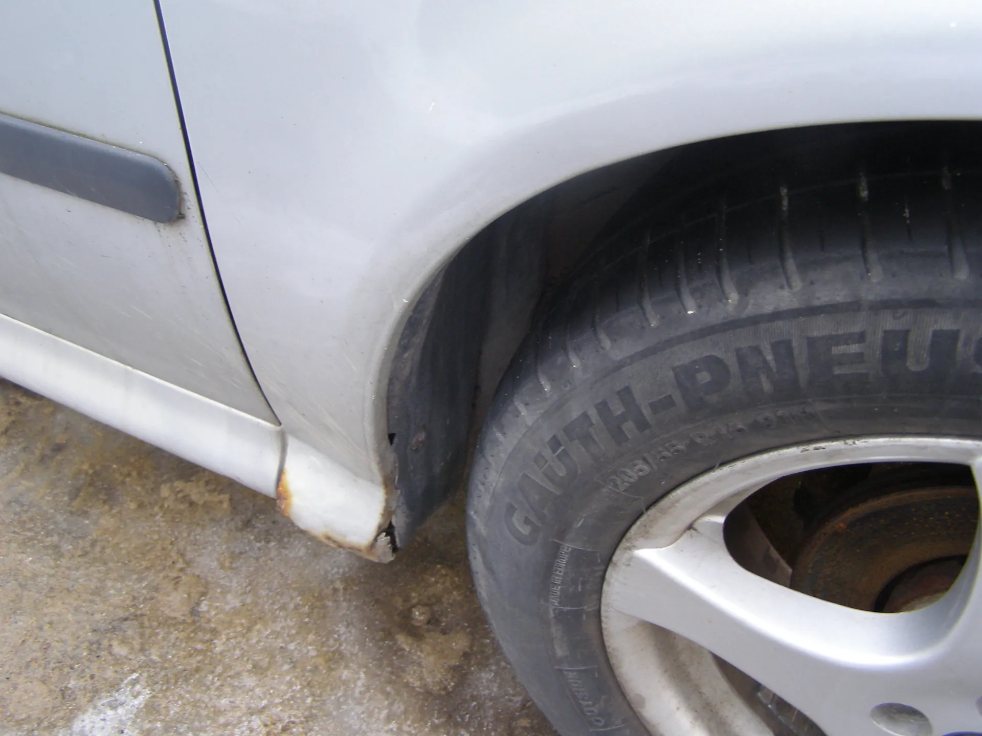 Zbliżenie na przednie koło samochodu w kolorze srebrnym. Widoczna opona z napisem GAUTH-PNEU oraz oznaczeniami 205/55 R16 91V. Felga aluminiowa w kolorze srebrnym, z widocznym rdzewiejącym dyskiem hamulcowym. Nadkole ma uszkodzenia i ślady korozji w dolnej części. Tło stanowi betonowa powierzchnia z plamami brudu.