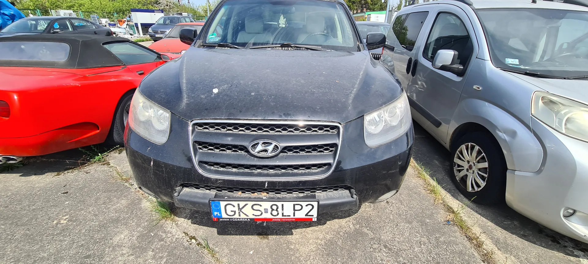Czarny samochód marki Hyundai z tablicą rejestracyjną GKS-8LP2 (PL, napisy 'jestem z GDANSKA' i 'jedź z rozwagą' pod tablicą) zaparkowany na placu parkingowym. Po lewej stronie czerwony kabriolet, po prawej srebrny samochód osobowy. W tle inne pojazdy, budynki i zieleń. Samochody stoją na betonowej nawierzchni z widoczną trawą w szczelinach.