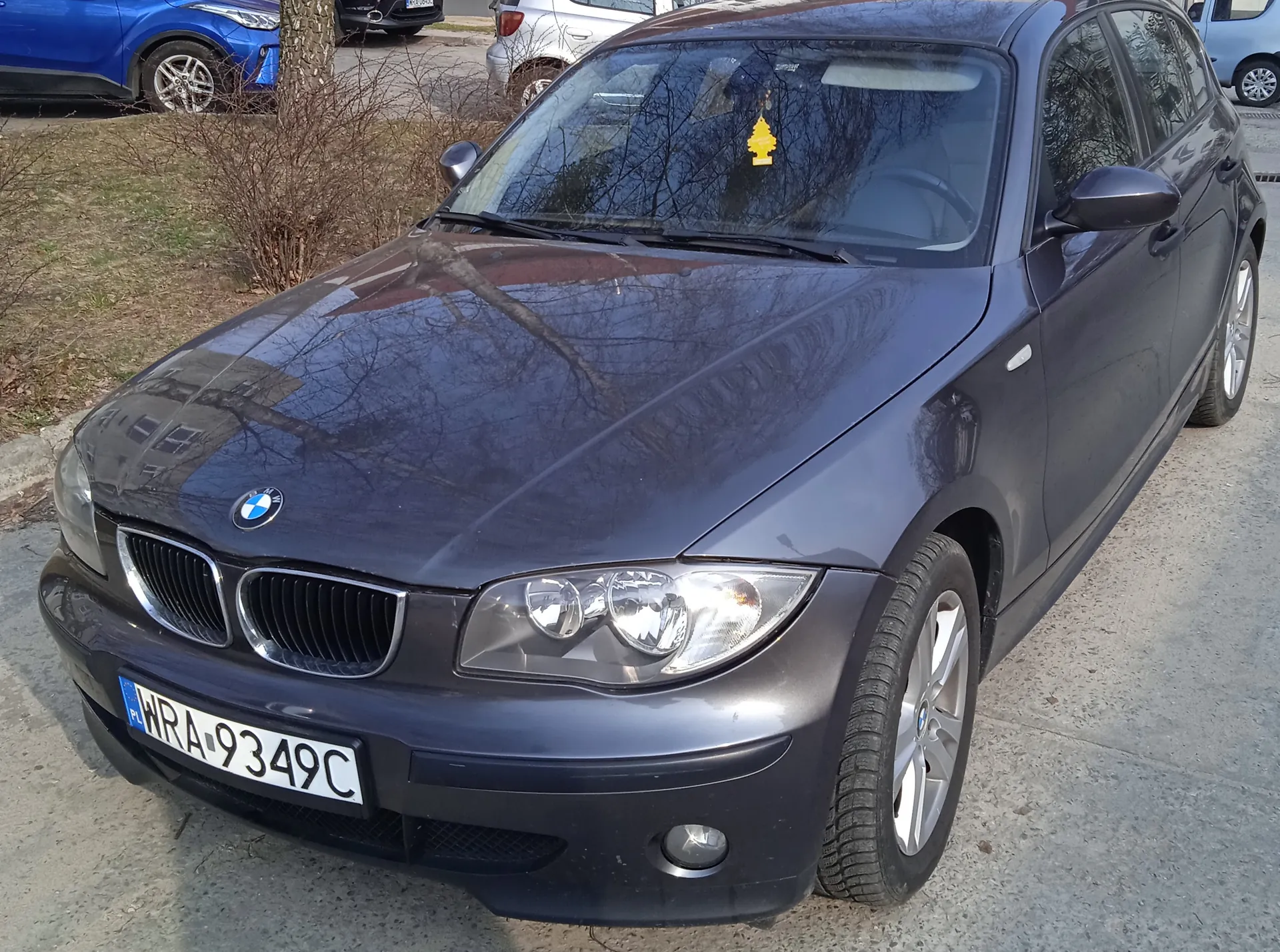 Ciemnoszary samochód marki BMW zaparkowany na betonowej powierzchni. Widoczna tablica rejestracyjna z napisem WRA-9349C oraz niebieskim polskim znakiem PL po lewej stronie. Na przedniej szybie wiszący żółty zapachowy w kształcie drzewka. W tle inne pojazdy: niebieski samochód, srebrny samochód i jasnoniebieski van. Wokół sucha trawa, krzewy i drzewo bez liści. Samochód ma charakterystyczne podwójne reflektory i klasyczną atrapę chłodnicy BMW.