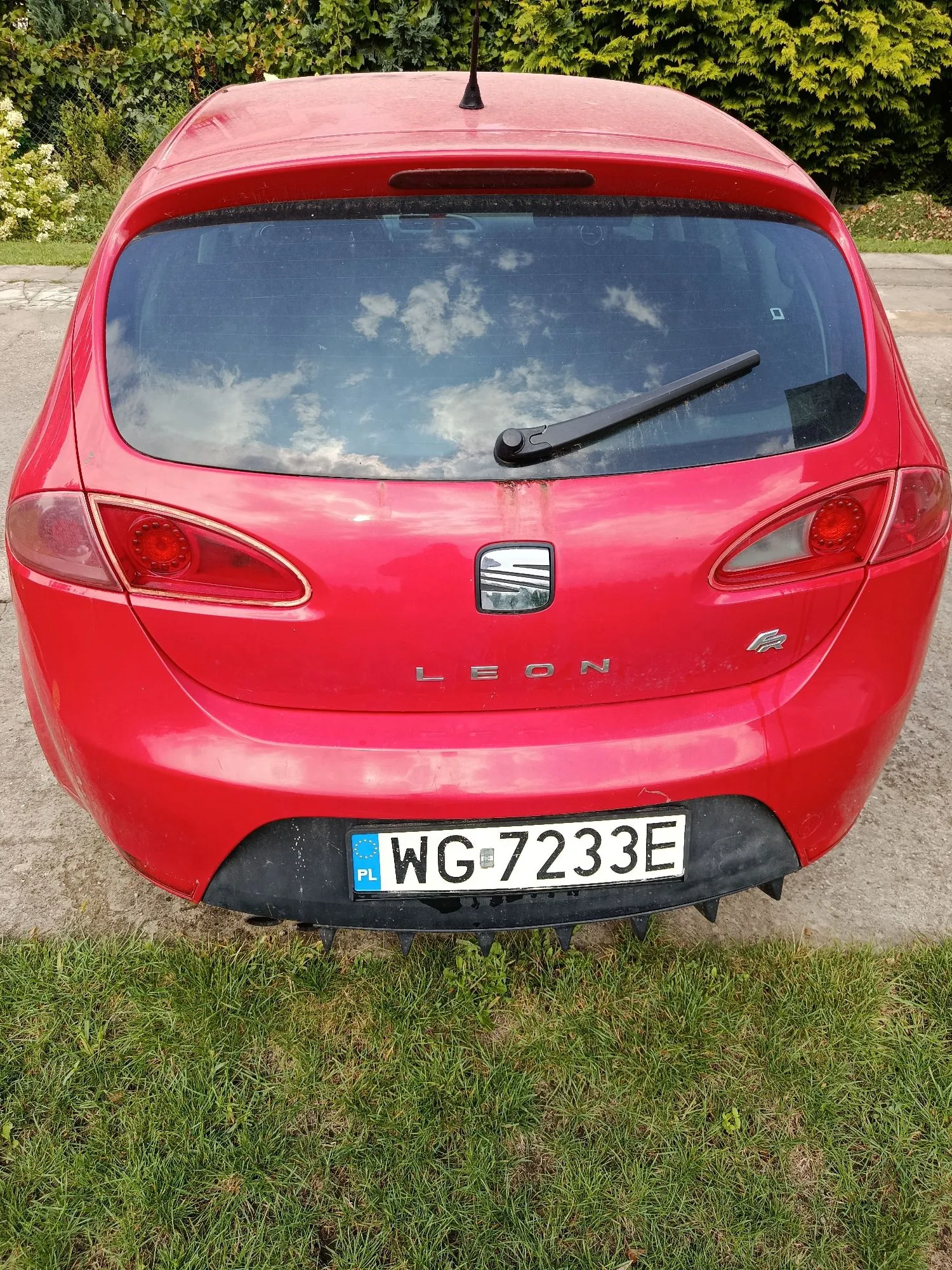 Tylny widok czerwonego samochodu marki Seat Leon. Na klapie bagażnika znajduje się logo marki, napis 'LEON' oraz oznaczenie 'FR'. Samochód posiada polską tablicę rejestracyjną o numerze WG 7233E. Pojazd stoi na trawie na tle zielonych krzewów.