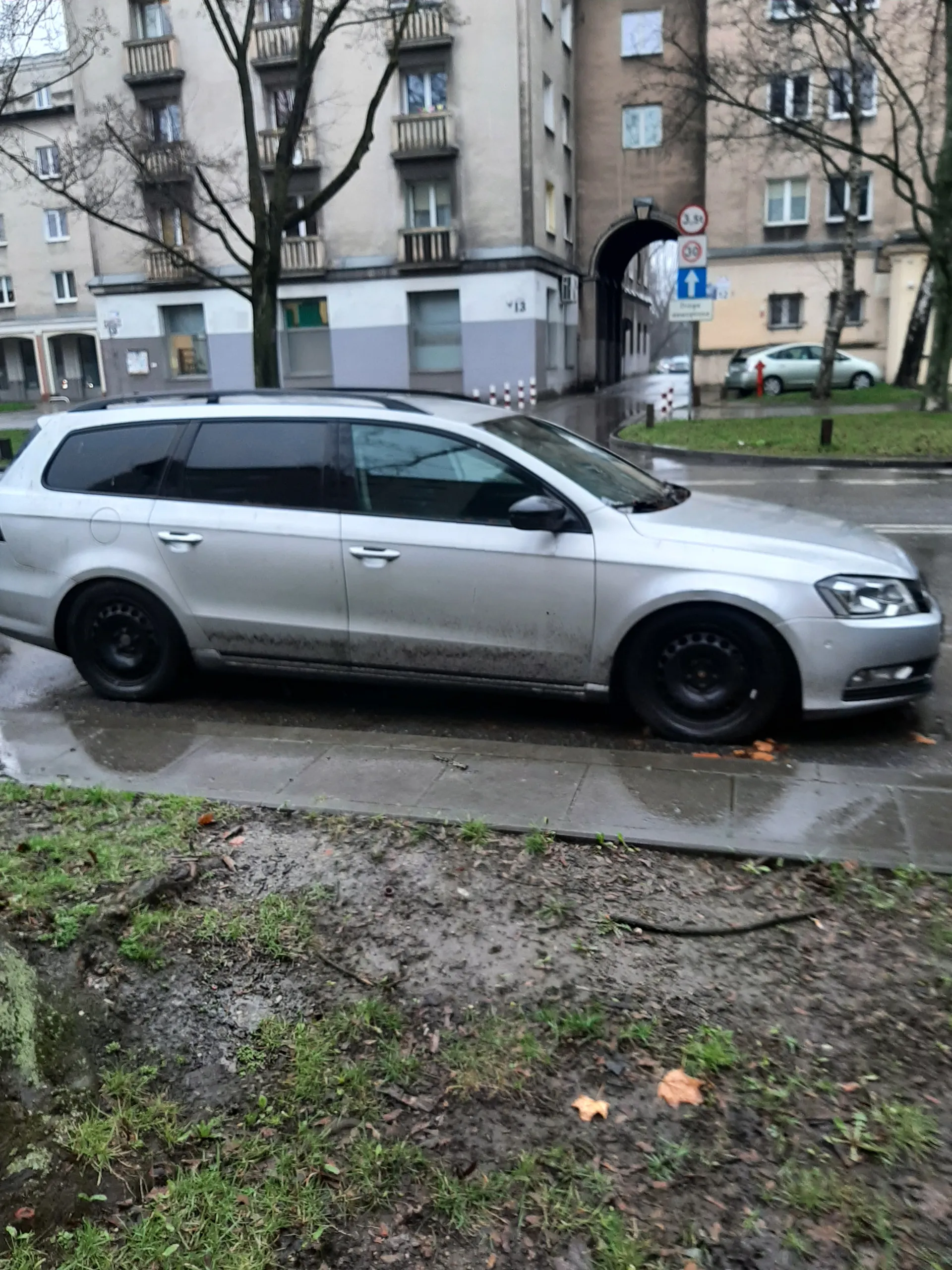 Srebrny samochód osobowy zaparkowany przy chodniku. Samochód ma czarne felgi i ciemne szyby. Po prawej stronie chodnik, a obok niego obszar z trawą i suchymi liśćmi. W tle budynki mieszkalne o różnych kolorach z oknami. Widoczne drogowskazy: niebieski znak z białą strzałką w prawo, czerwony okrągły znak oraz inny znak. Po lewej drzewa bez liści. Droga wydaje się mokra.