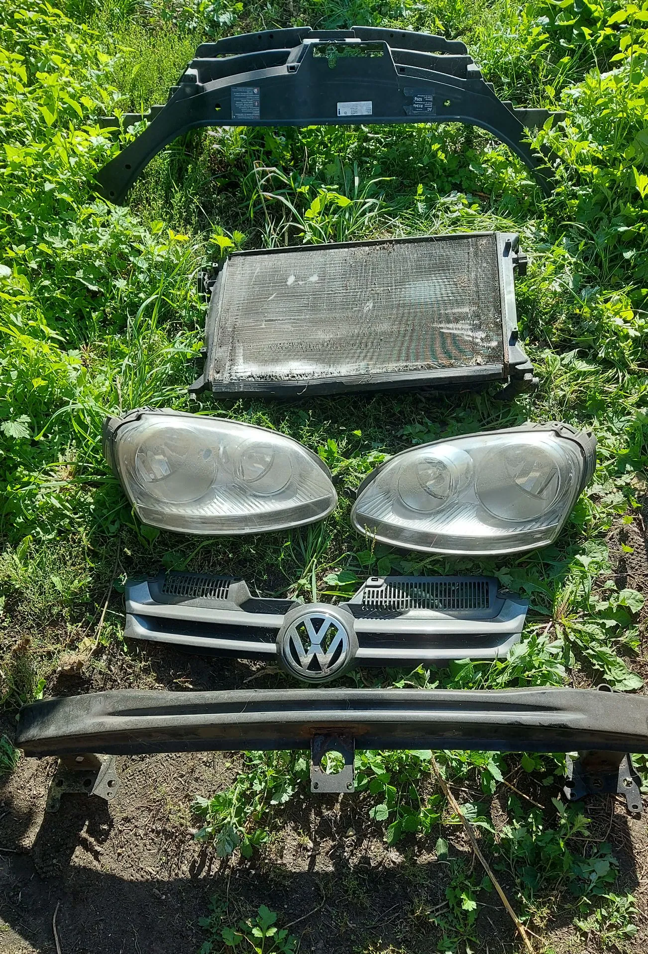Zestaw części samochodowych rozłożonych na zielonej trawie. Od góry: czarny plastikowy wzmocniony panel przedni, chłodnica, dwa reflektory przednie, srebrna atrapa chłodnicy z logo Volkswagena oraz czarna belka zderzaka.