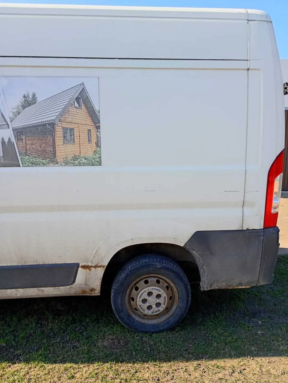 Biały furgon zaparkowany na trawie. Na boku furgonu znajduje się zdjęcie drewnianego domku z dachem krytym dachówką. Widoczne są tylne koło z brudnym felgiem, zarys zardzewiałych plam pod kołem oraz czerwone światło tylnego reflektora. Tło stanowi niebieskie niebo i fragment brązowej konstrukcji po prawej stronie. Trawa pod furgonem jest częściowo wysuszona.