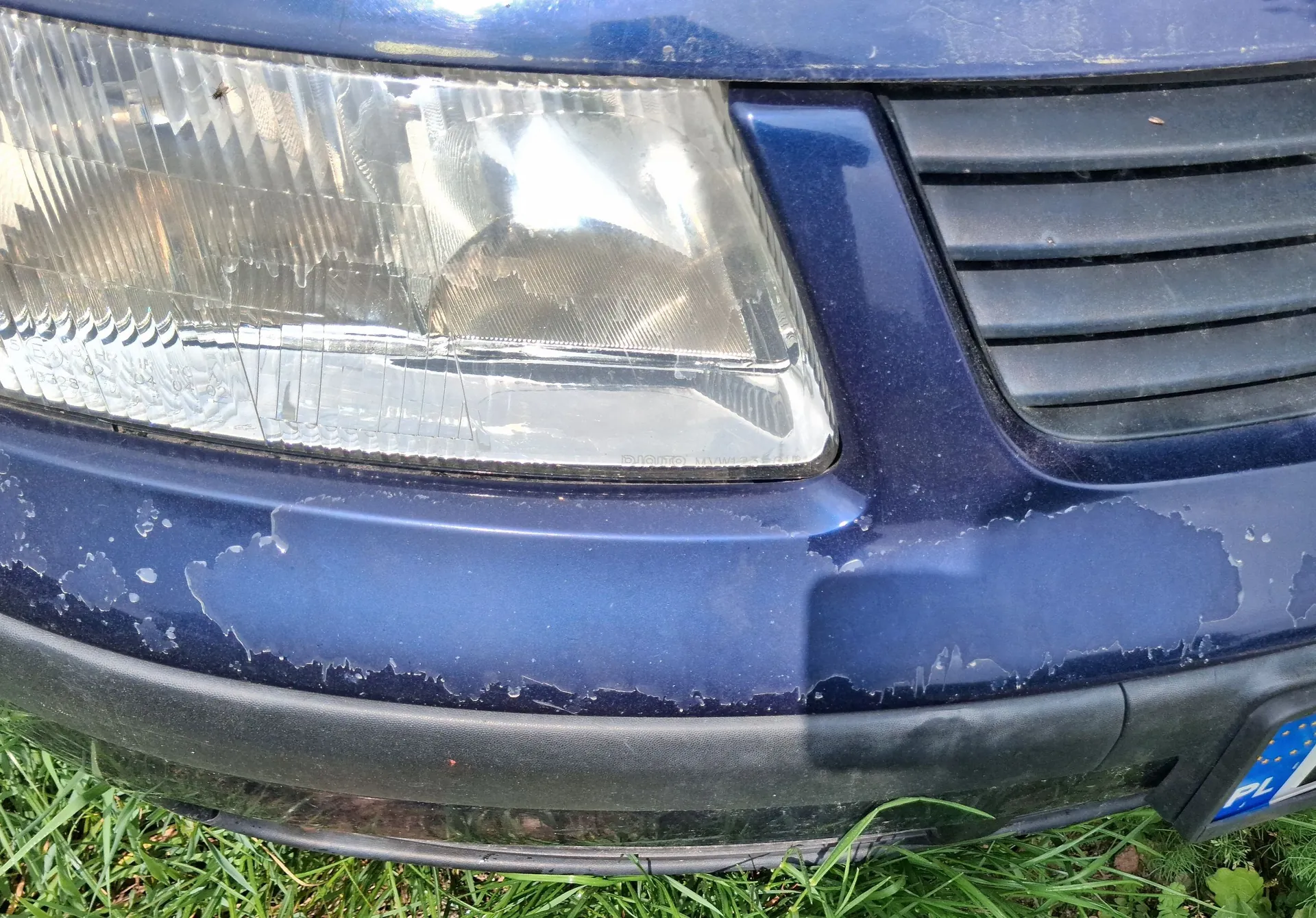 Zbliżenie na przednią część ciemnoniebieskiego samochodu. Widoczny jest przedni reflektor oraz fragment grilla. Lakier na zderzaku pod reflektorem jest mocno zniszczony, z widocznymi odpryskami i łuszczącą się powłoką. Samochód stoi na zielonej trawie.