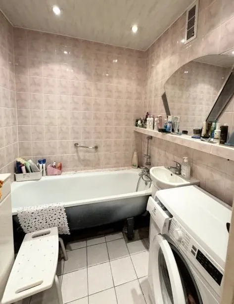 Wnętrze łazienki z różowymi płytkami na ścianach i białymi płytkami na podłodze. W pomieszczeniu znajduje się biała wanna z ciemnym podstawem, umywalka, pralka oraz półka z różnymi kosmetykami i butelkami. Na krawędzi wanny leży biały ręcznik z wzorem. Obok wanny stoi biały stołek. Sufit wyposażony jest w wewnętrzne oświetlenie, a na ścianie widoczne jest lustro oraz wentylacja.