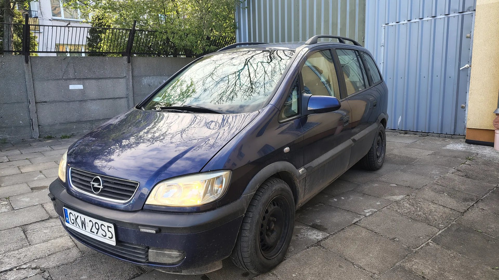 Ciemnoniebieski samochód Opel z tablicą rejestracyjną GKW 29S5 zaparkowany na brukowanej powierzchni. W tle betonowy mur z metalowym ogrodzeniem, drzewa, budynek z oknami oraz niebieska metalowa brama. Samochód posiada dachowy bagażnik i widoczne zabrudzenia na dolnej części nadwozia.