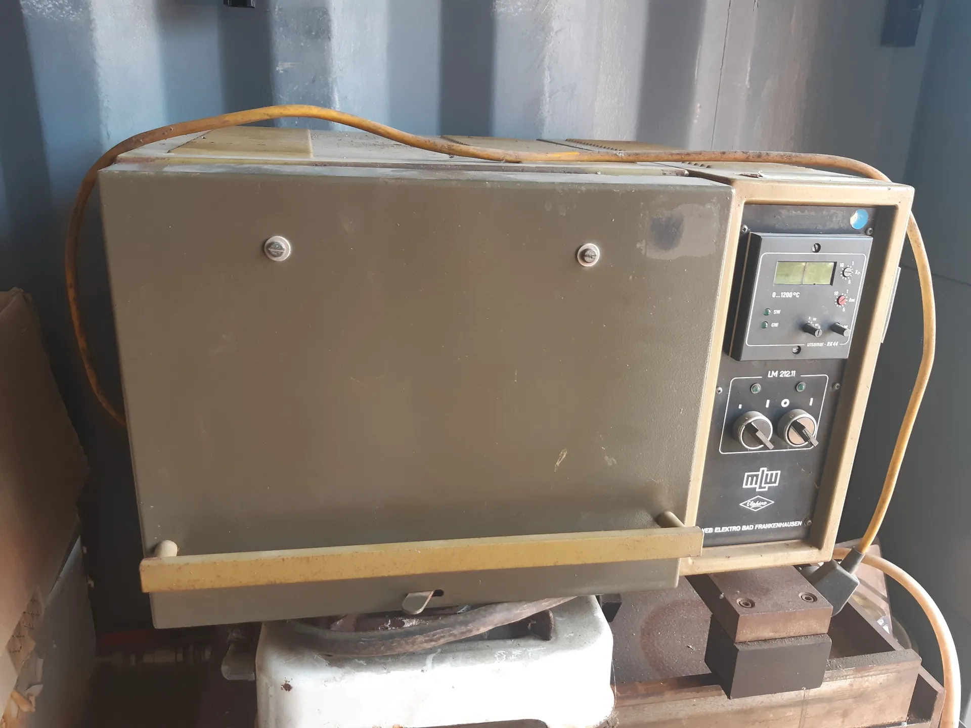Oliwkowy, metalowy aparat pomiarowy lub sterownik przemysłowy z panelem kontrolnym po prawej stronie. Panel zawiera cyfrowy wyświetlacz z wartością '1200°C', kilka przełączników, pokrętło oraz logo 'mlw'. Urządzenie jest połączone z żółtym kablem zasilającym, który biegnie nad jego obudową. Całość spoczywa na białym podłożu w pomieszczeniu o szarych, metalowych ścianach.