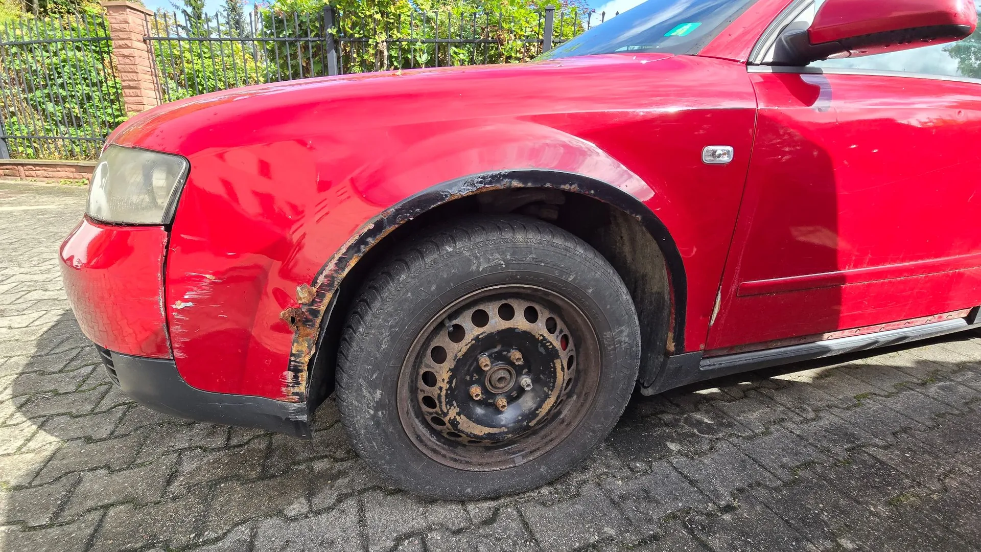 Zbliżenie na przedni lewy błotnik czerwonego samochodu. Nad kołem widoczne są znaczne uszkodzenia karoserii w postaci rdzy i ubytków lakieru. Samochód posiada czarną stalową felgę i jest zaparkowany na kostce brukowej. W tle widać zielony płot i roślinność.