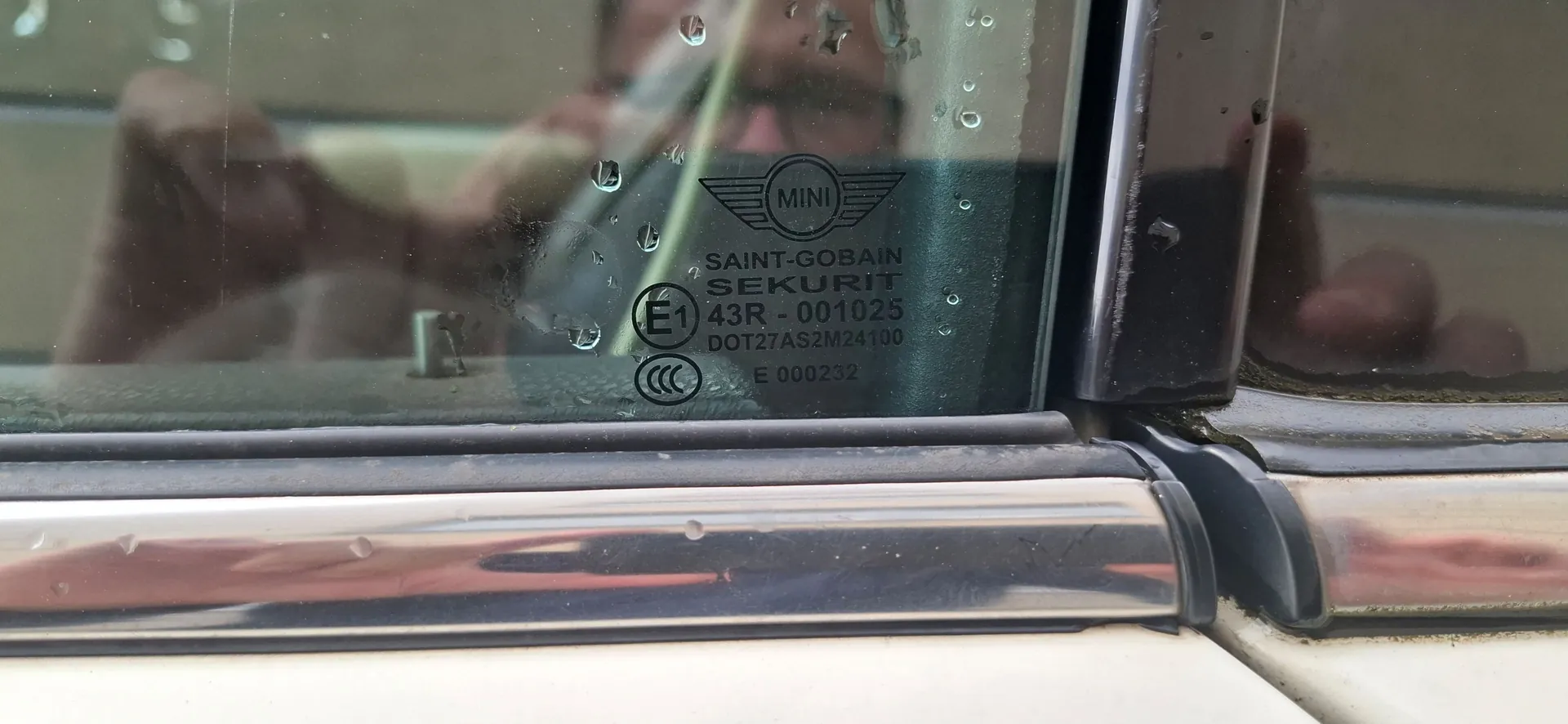 Zbliżenie na szybę samochód marki Mini z widocznymi kroplami wody. Na szybie znajduje się grawer z logo MINI oraz tekstem: 'SAINT-GOBAIN SEKURIT', 'E1 43R - 001025', 'DOT27AS2M24100' oraz 'E 000232'. Poniżej szyby widoczna jest chromowana listwa wykończeniowa.