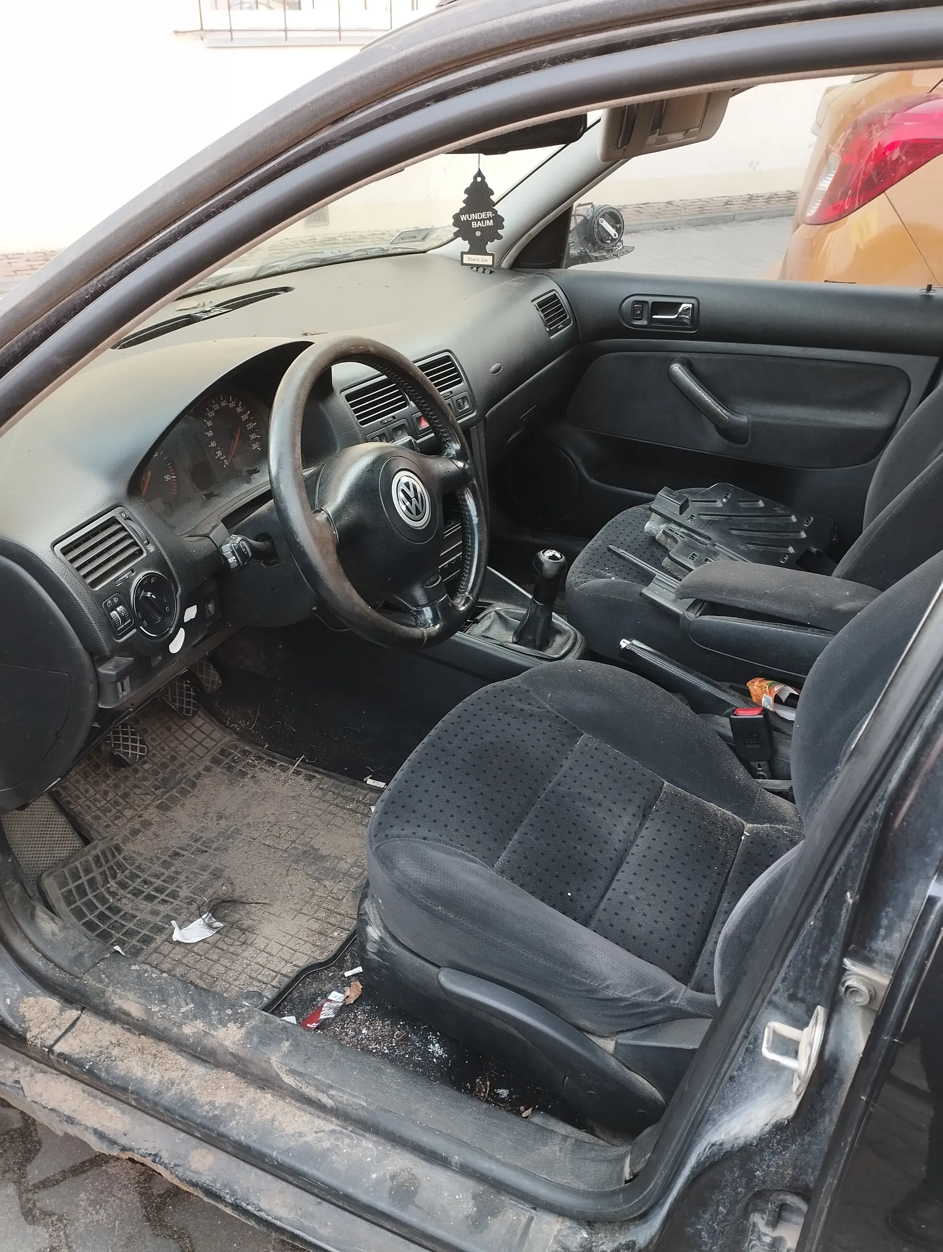 Wnętrze samochodu marki Volkswagen z otwartą drzwią kierowcy. Widoczne czarne siedzenia z kropkowanym wzorem, kierownica z logo VW, zanieczyszczony dywanik podłogowy z resztkami śmieci, deska rozdzielcza z licznikami oraz wiszący na lusterku wstecznym odświeżacz powietrza w kształcie choinki z napisem "WUNDER-BAUM". Po prawej stronie widoczny fragment innego pojazdu o pomarańczowej karoserii. Wnętrze samochodu jest zabrudzone, a na siedzeniu pasażera leżą rozrzucone przedmioty.