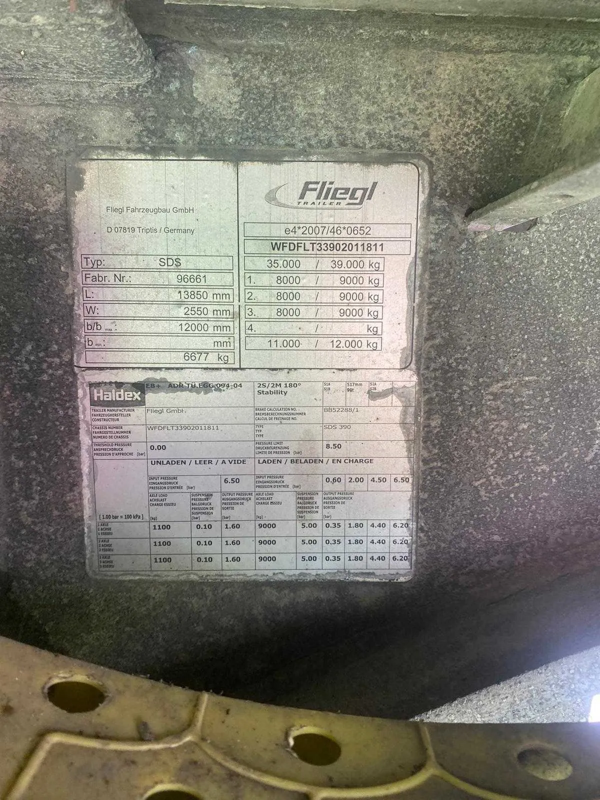 Zdjęcie dwóch tablic znamionowych przymocowanych do metalowej powierzchni pojazdu. Górna tablica firmy Fliegl zawiera dane techniczne: Typ SDS, Fabr. Nr. 96661, wymiary L: 13850 mm, W: 2550 mm, b/b: 12000 mm oraz masę całkowitą 39,000 kg. Dolna tablica firmy Haldex dotyczy układu hamulcowego, zawiera numer WFDPLT33902011811 oraz tabelę z wartościami ciśnień i obciążeń dla różnych osi.