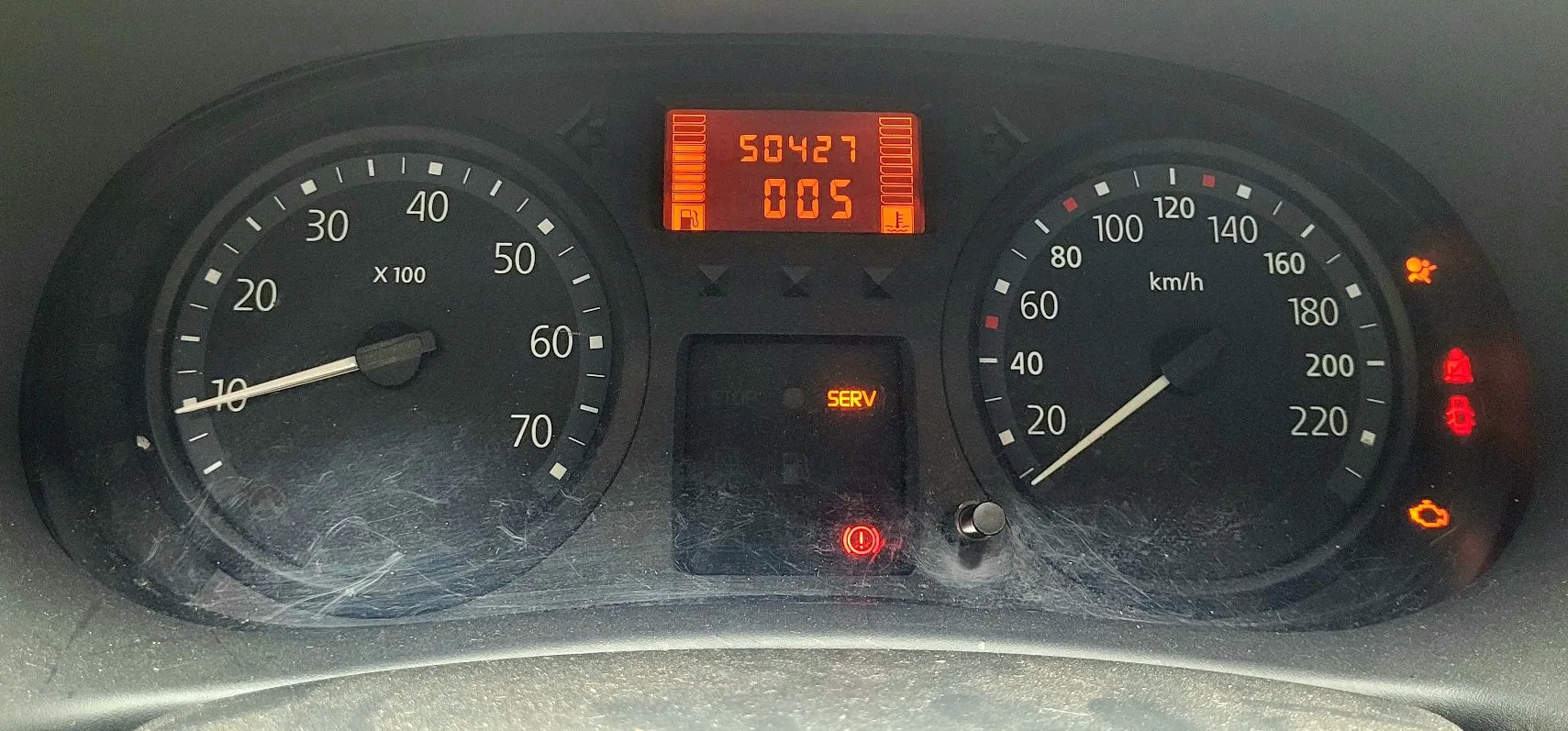 Zbliżenie na deskę rozdzielczą samochodu. Po lewej stronie znajduje się wskaźnik paliwa i temperatury, po prawej prędkościomierz z zaznaczeniami od 20 do 220 km/h. W górnej części znajduje się cyfrowy wyświetlacz z liczbami 50427 oraz 005. Na środku widnieje napis SERV oraz czerwona kontrolka ostrzegawcza z wykrzyknikiem. Po prawej stronie prędkościomierza świecą trzy pomarańczowe i czerwone kontrolki ostrzegawcze.