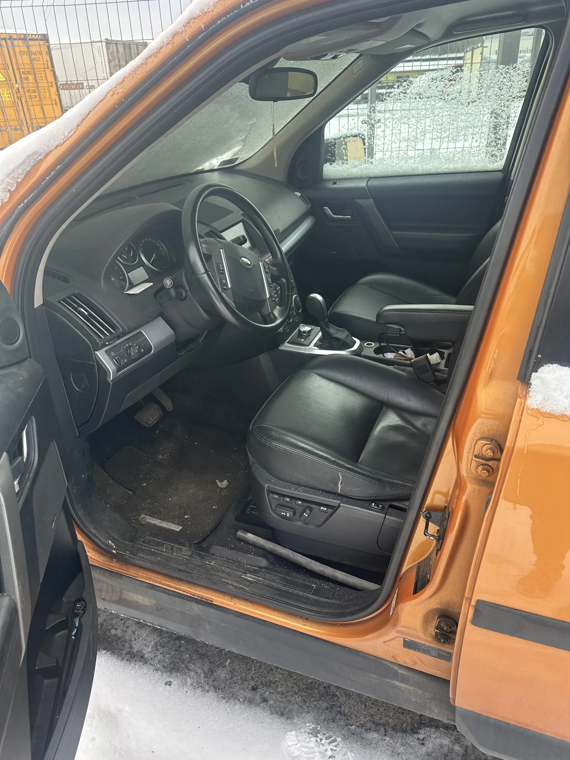 Wnętrze pomarańczowego samochodu z otwartą przodem drzwiami kierowcy. Widoczne czarne skórzane fotele, kierownica z logo, deska rozdzielcza z wskaźnikami oraz przyciskami. Na podłodze brudne dywaniki, a na siedzeniu pasażera leży metalowy pręt. Poza samochodem śnieg na ziemi i na dachu pojazdu, metalowe ogrodzenie oraz pomarańczowe kontenery w tle. Szkła samochodu częściowo pokryte szronem.