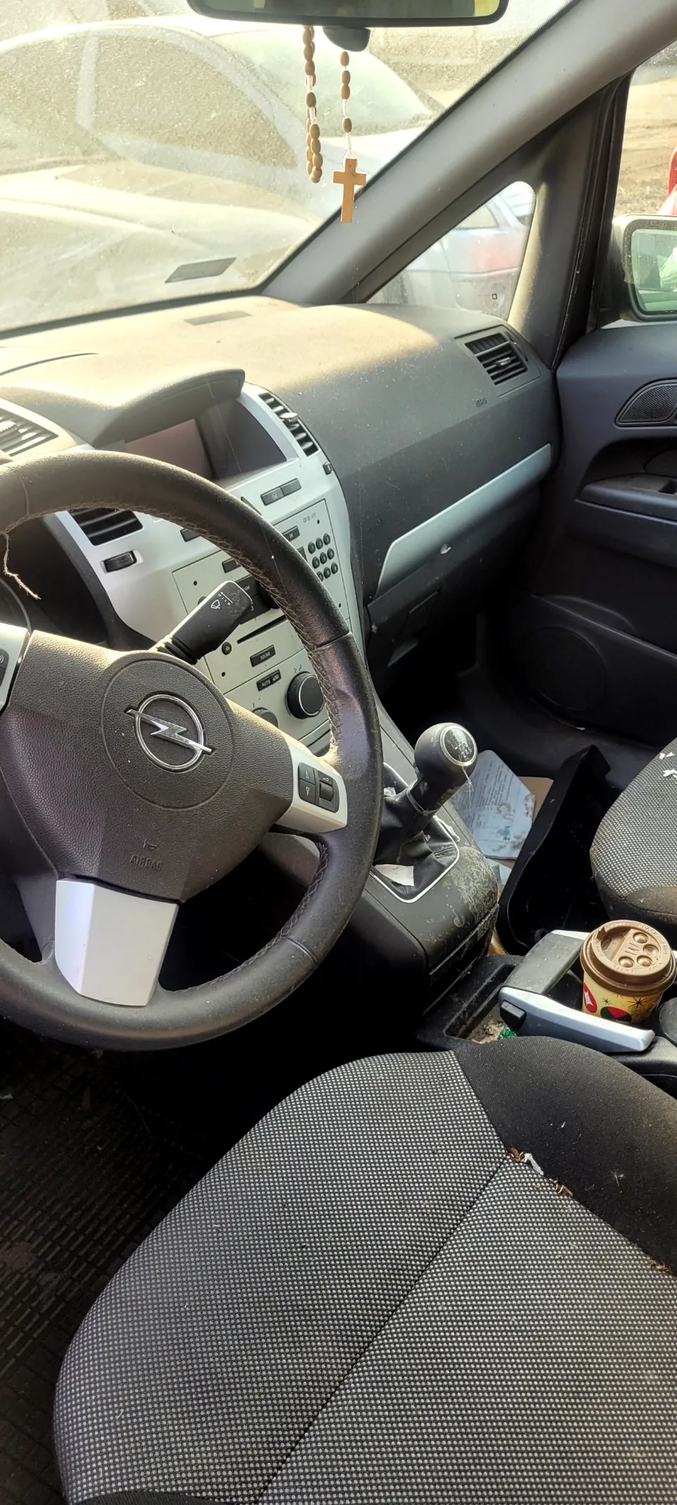 Wnętrze samochodu z kierownicą z logo Opel, deską rozdzielczą, dźwignią zmiany biegów, czarno-szarymi fotelami z kropkowanym wzorem, kubkiem kawy w uchwycie oraz różańcem z krzyżem wiszącym na lusterku wstecznym. Przez przednią szybę widoczny jest inny samochód.