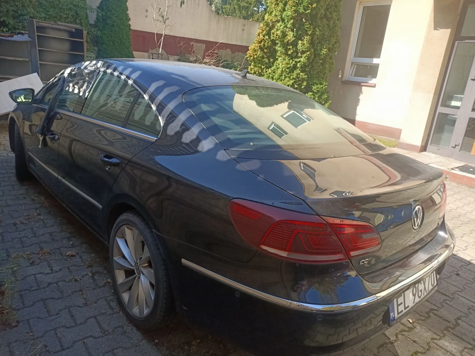 Czarny samochód marki Volkswagen CC zaparkowany na kostce brukowej. Zdjęcie przedstawia tył i prawy bok pojazdu. W tle widoczny jest fragment budynku z oknem oraz zielone krzewy. Na tablicy rejestracyjnej widnieje napis EL 9GX0.