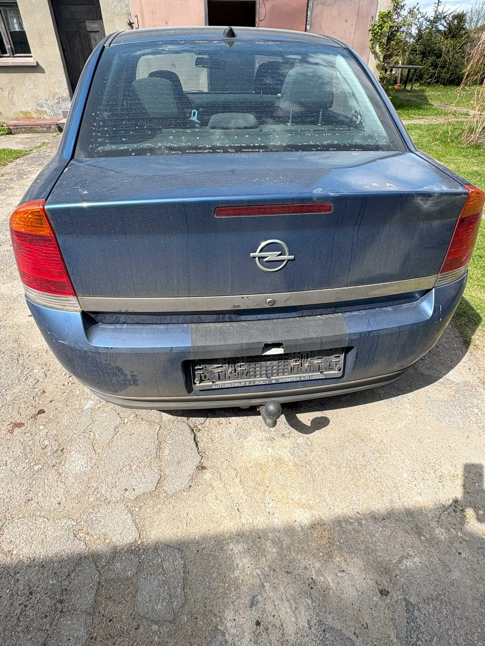 Z tyłu niebieski samochód marki Opel z widocznym logo na bagażniku. Na środku bagażnika znajduje się czerwony pasek przeciwmgielny. Tablica rejestracyjna jest częściowo zakryta czarną taśmą, pod nią widoczny napis: 'AUTOBEDRIJF E.J. ROOY • KRIMPEN A/D IJSSEL'. Samochód stoi na betonowej powierzchni, w tle budynek z drzwiami i oknem oraz zieleń. Bagażnik i szyba są lekko zabrudzone.