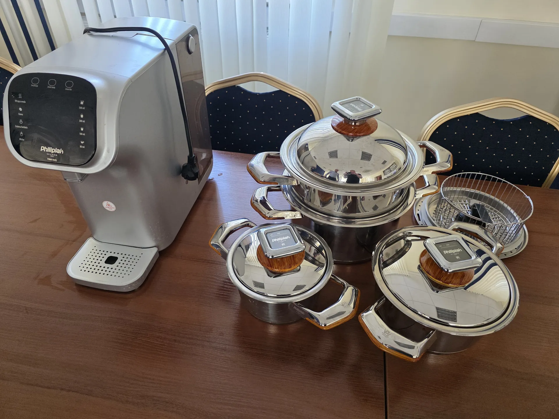 Na drewnianym stole umieszczono srebrny podgrzewacz wody marki Phillipak z czarnym panelem sterowania i widocznymi napisami: 'Phillipak', '500 ml', '300 ml', '180 ml', '80 ml'. Obok znajdują się trzy srebrne garnki z pokrywkami marki Phillipak, z drewnianymi uchwytami na pokrywkach oraz metalowy koszyk. W tle widoczne są krzesła z ciemnoniebieską tapicerką w złote kropki i białe żaluzje.