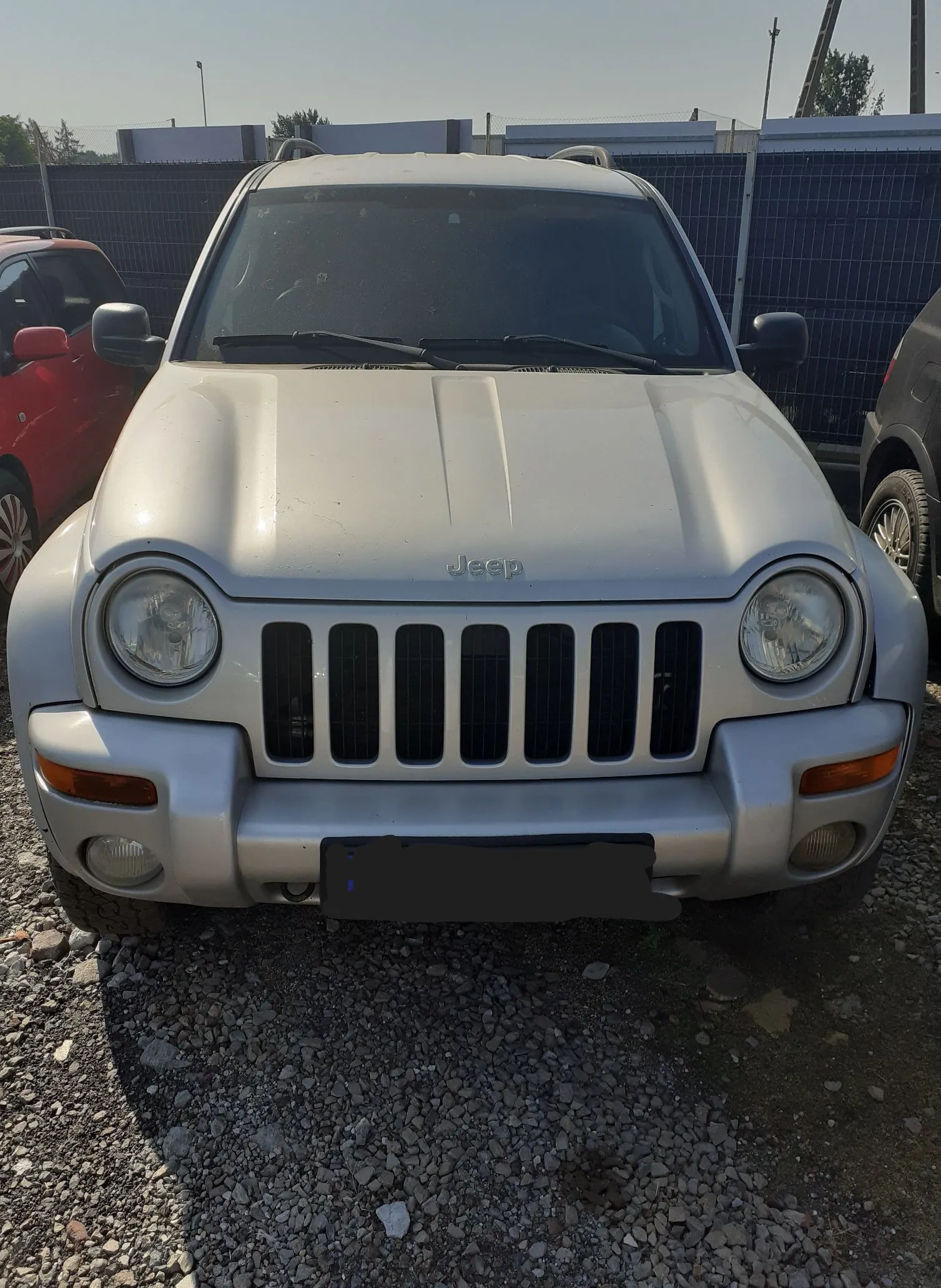 Srebrny samochód marki Jeep widziany od przodu. Widoczny jest charakterystyczny grill z siedmioma pionowymi szczelinami, okrągłe reflektory oraz napis 'Jeep' na masce. Auto stoi na żwirowym podłożu, a po lewej stronie widoczny jest fragment czerwonego samochodu.