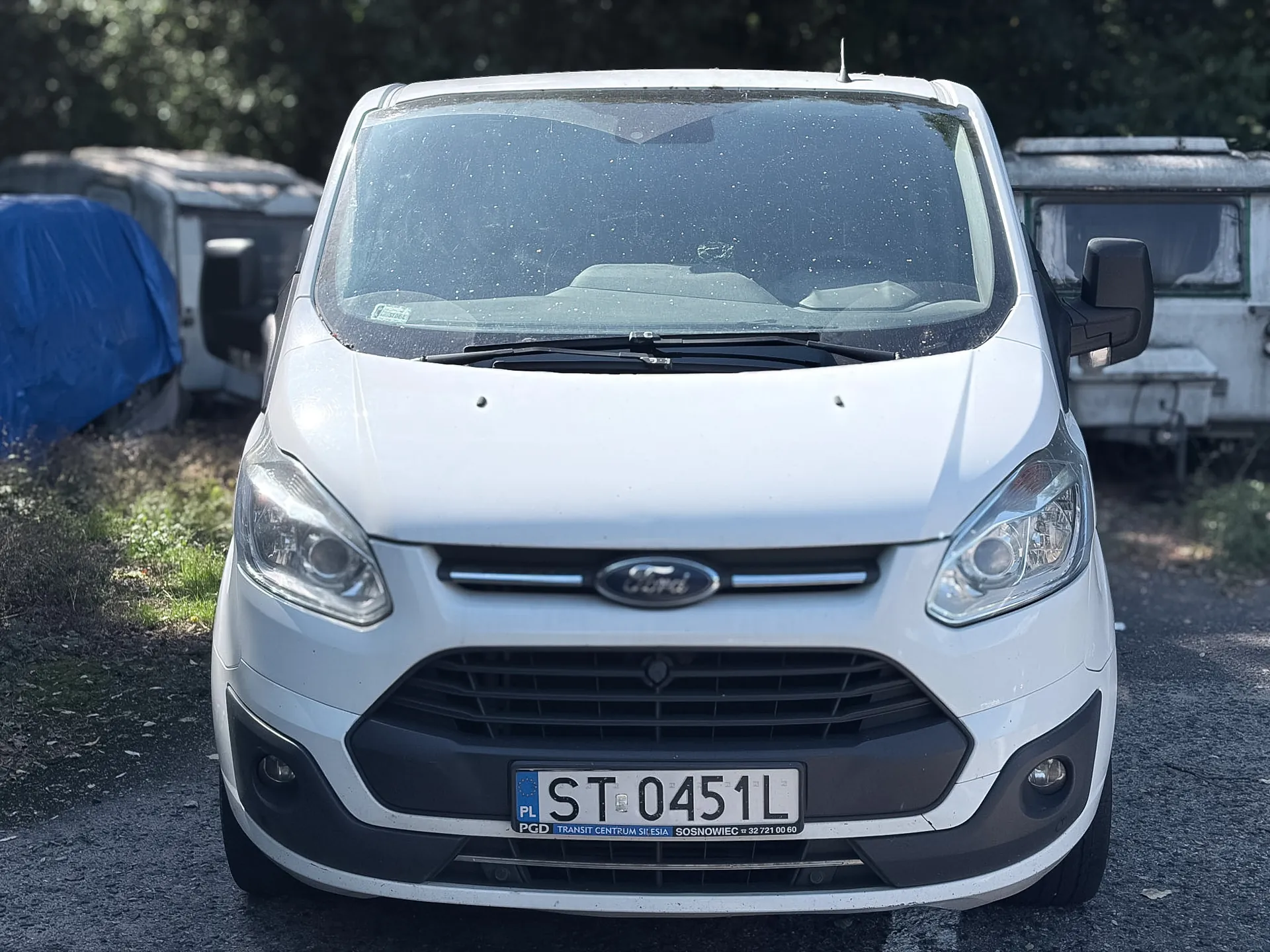 Widok z przodu białego vana marki Ford. Pojazd ma zabrudzoną przednią szybę i tablicę rejestracyjną o numerze ST 0451L. Poniżej numeru rejestracyjnego znajduje się napis: PGD TRANSIT CENTRUM SK ESIA SOSNOWIEC tel. 32 721 00 60. W tle widoczne są inne pojazdy, w tym jeden przykryty niebieską plandeką.