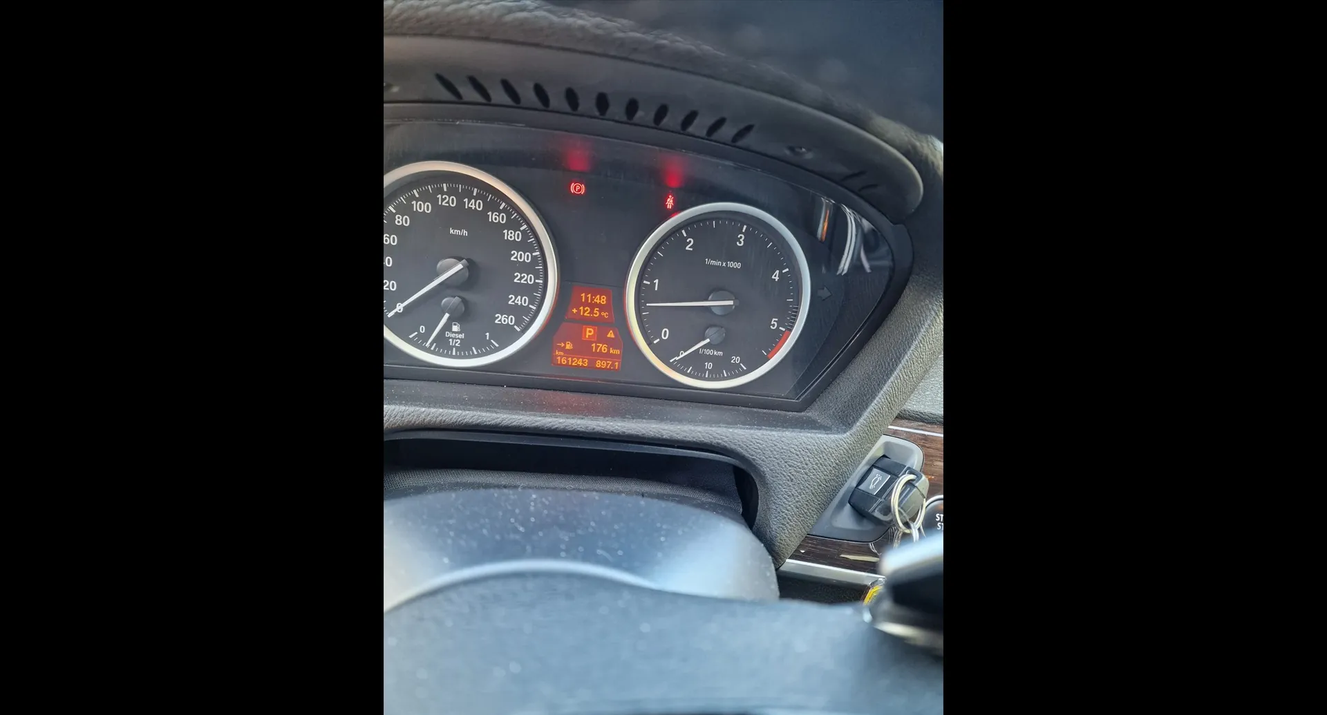 Zbliżenie na deskę rozdzielczą samochodu. Widoczne są dwa główne analogowe wskaźniki: prędkościomierz po lewej stronie (skala do 260 km/h) oraz obrotomierz po prawej stronie (skala do 5 tys. obr./min). Pomiędzy nimi znajduje się cyfrowy wyświetlacz pokazujący godzinę 11:48, temperaturę +12.5°C, włączony bieg P oraz przebieg 176 km i 807.1. Nad wskaźnikami świecą czerwone kontrolki ostrzegawcze. W dolnej części zdjęcia widoczny jest fragment kierownicy oraz kluczyki w stacyjce.